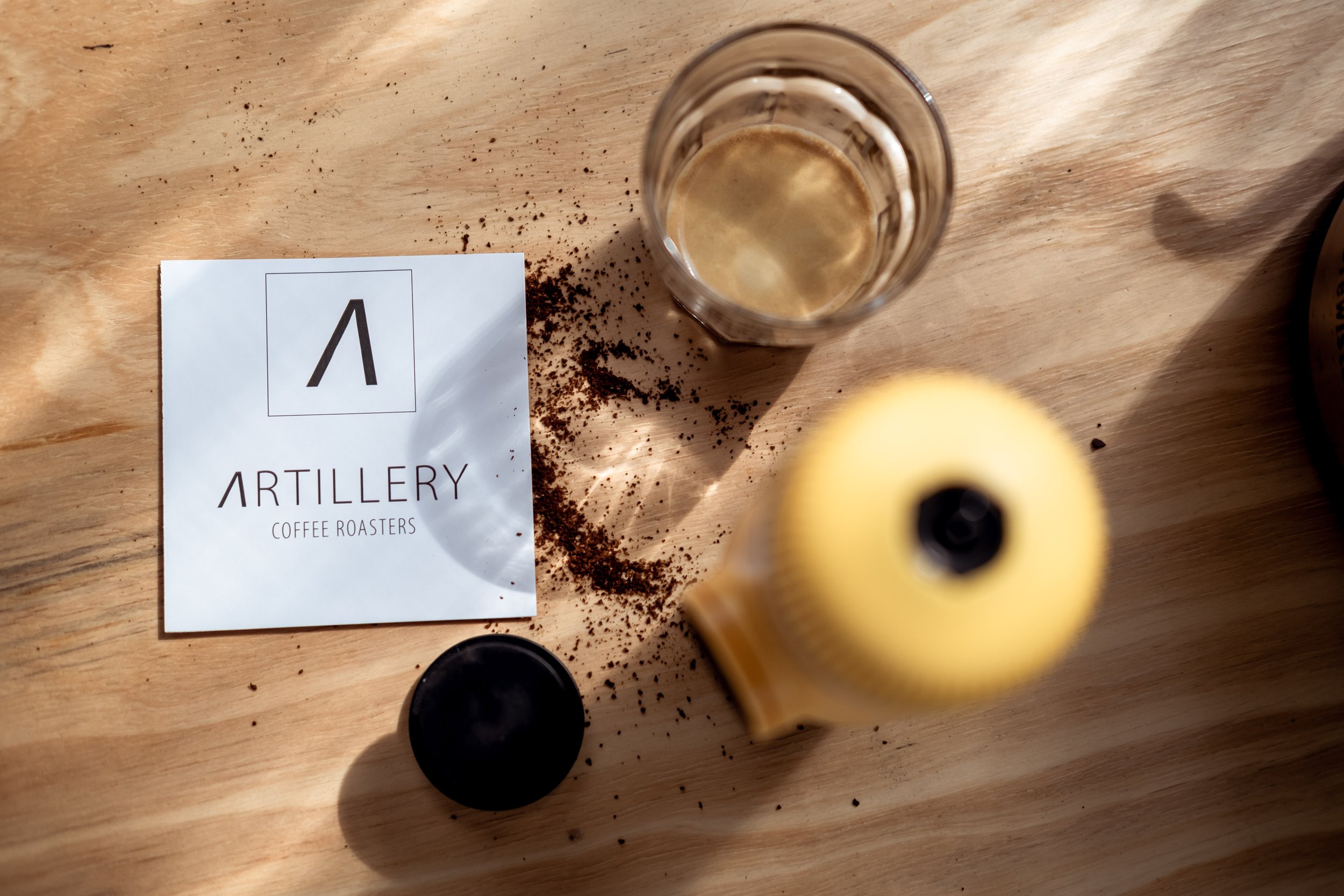 Artillery Coffee Roasters card on a wooden table with spilled coffee grounds, a glass of espresso, and a yellow coffee grinder.