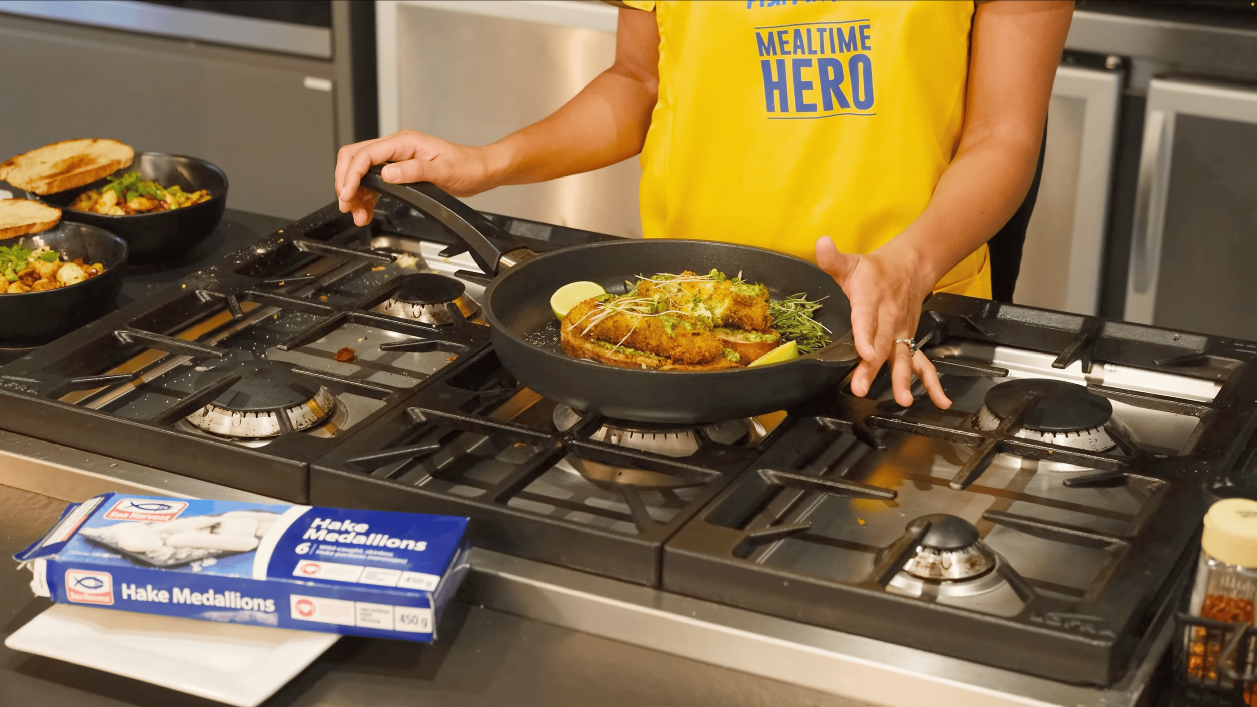 Person in a yellow apron placing a fried fish dish garnished with microgreens and lemon slices on a stove.