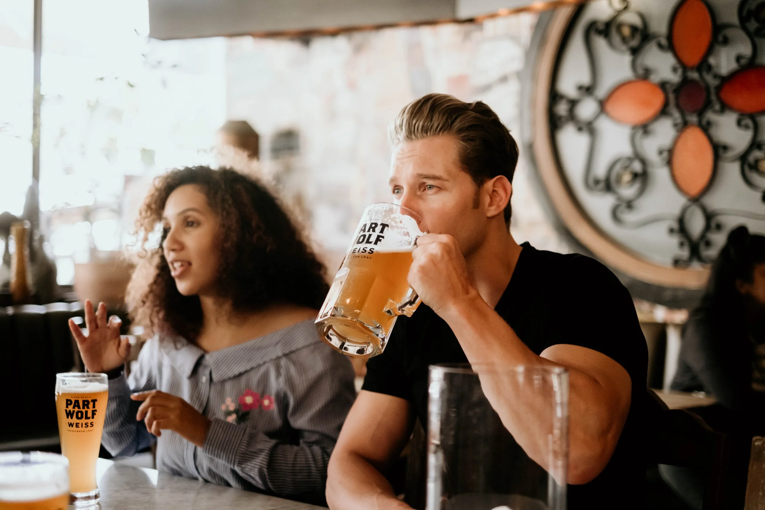 A man drinking beer at a bar with a woman sitting next to him, both having glasses of beer with 'PART WOLF WEISS' written on them. The man is taking a sip from his mug, while the woman is speaking and gesturing with her hand.