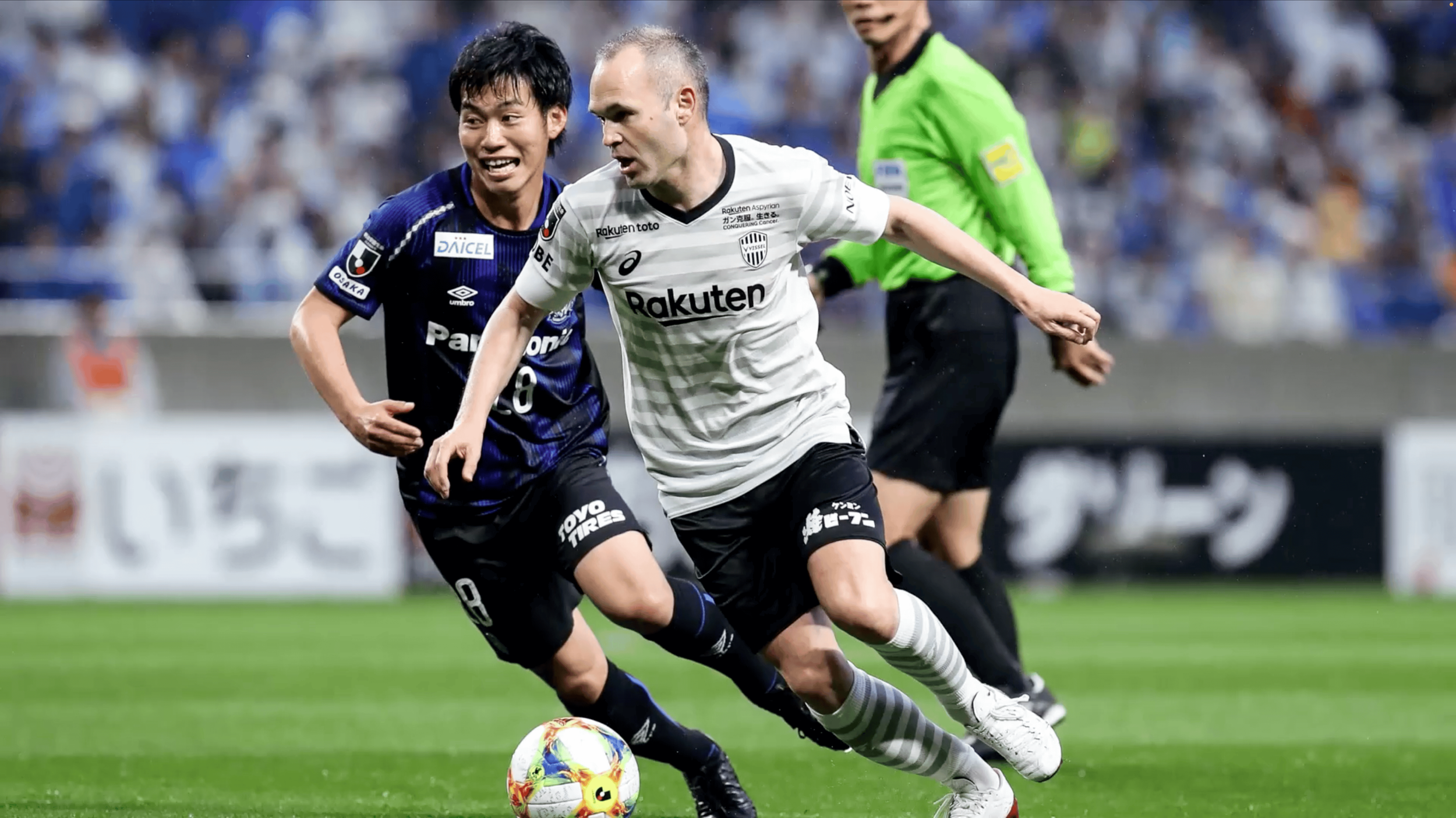 Two soccer players, one in a white and black uniform and the other in a blue and black uniform, chase a soccer ball on the field as an official watches in the background.