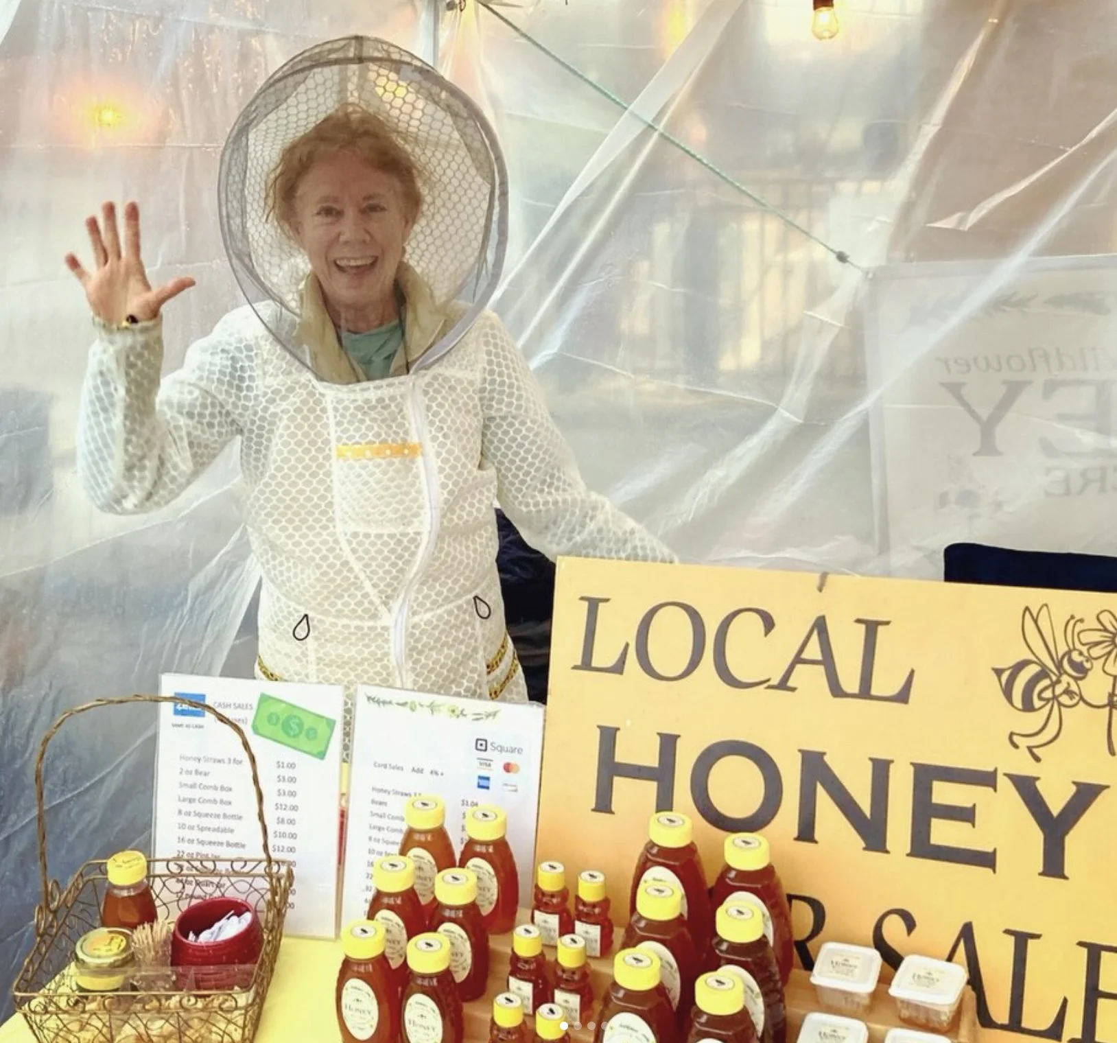 A cheerful woman wearing a white honeycomb-patterned jacket and a bee veil, standing behind a table at a local honey sale. The table displays bottles of honey, some packaged in small jars, with a yellow sign reading 'LOCAL HONEY SALE'. The woman is waving and smiling.