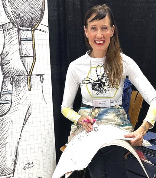 Person sitting and drawing on fabric, wearing a white shirt with an illustration and a name tag, with a large sketch on a board beside them.