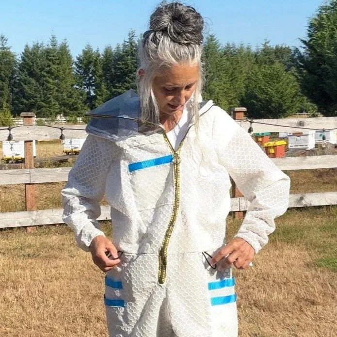 A woman with gray hair tied up, wearing a white protective suit with blue accents, standing outdoors in front of a wooden fence and trees.