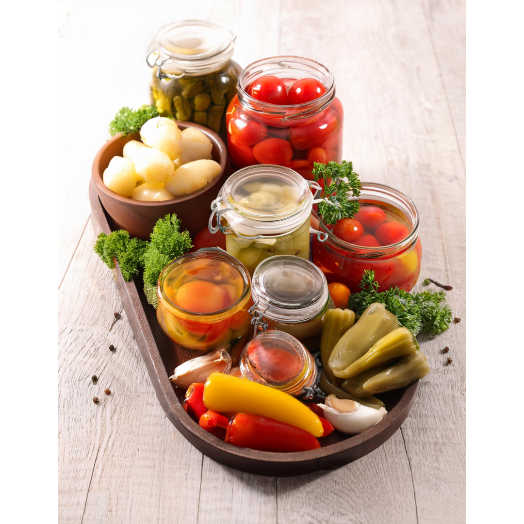 An assortment of preserved vegetables including cherry tomatoes, pickled peppers, garlic, and assorted condiments in glass jars on a wooden tray, garnished with parsley and black peppercorns.