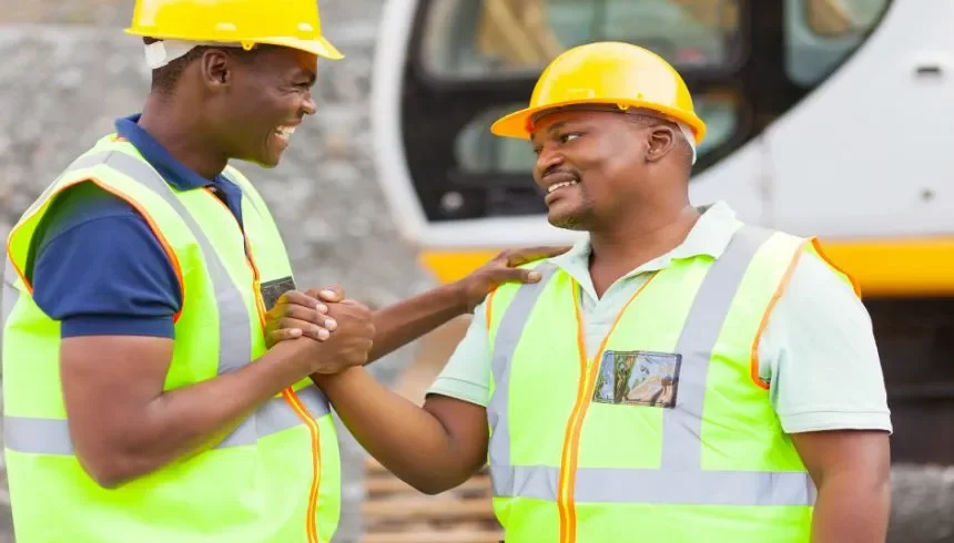 Two construction workers wearing yellow hard hats and reflective safety vests smiling and shaking hands at a construction site.