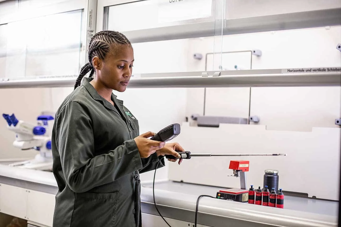 A female scientist in a lab coat looking at a handheld device, standing in a laboratory with scientific equipment and bottles on the counter.