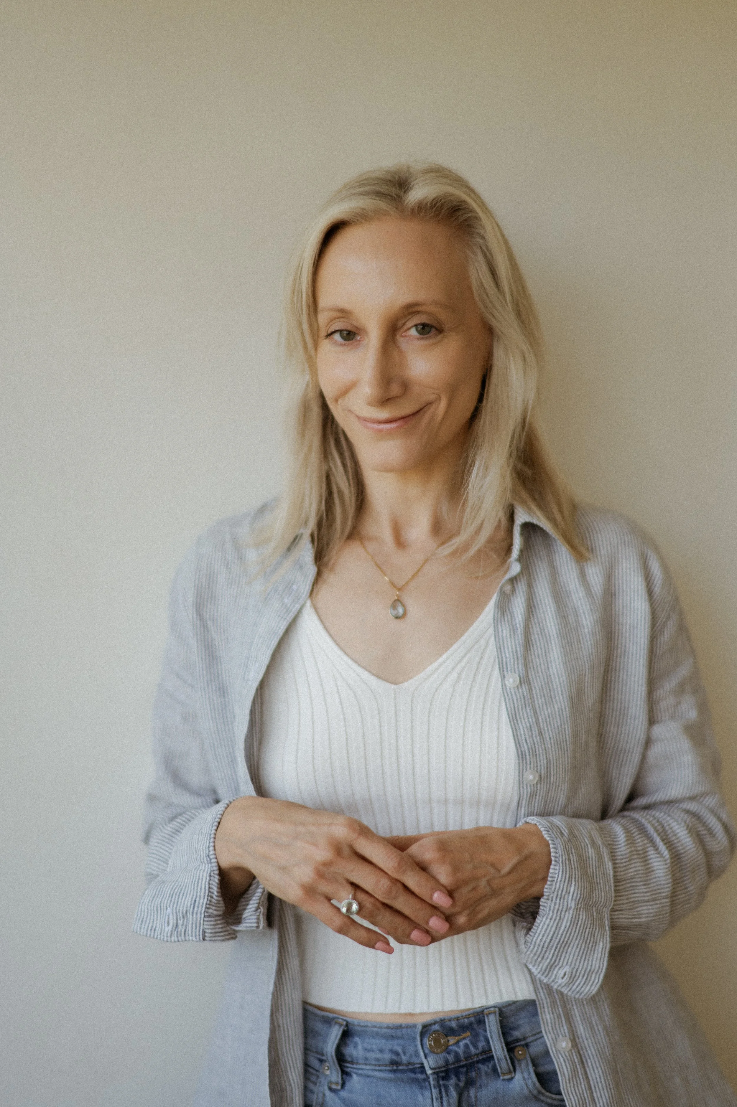 A woman with shoulder-length blonde hair smiling while looking at the camera, wearing a white ribbed top, a light blue striped shirt, and blue jeans, standing against a plain light-colored background.