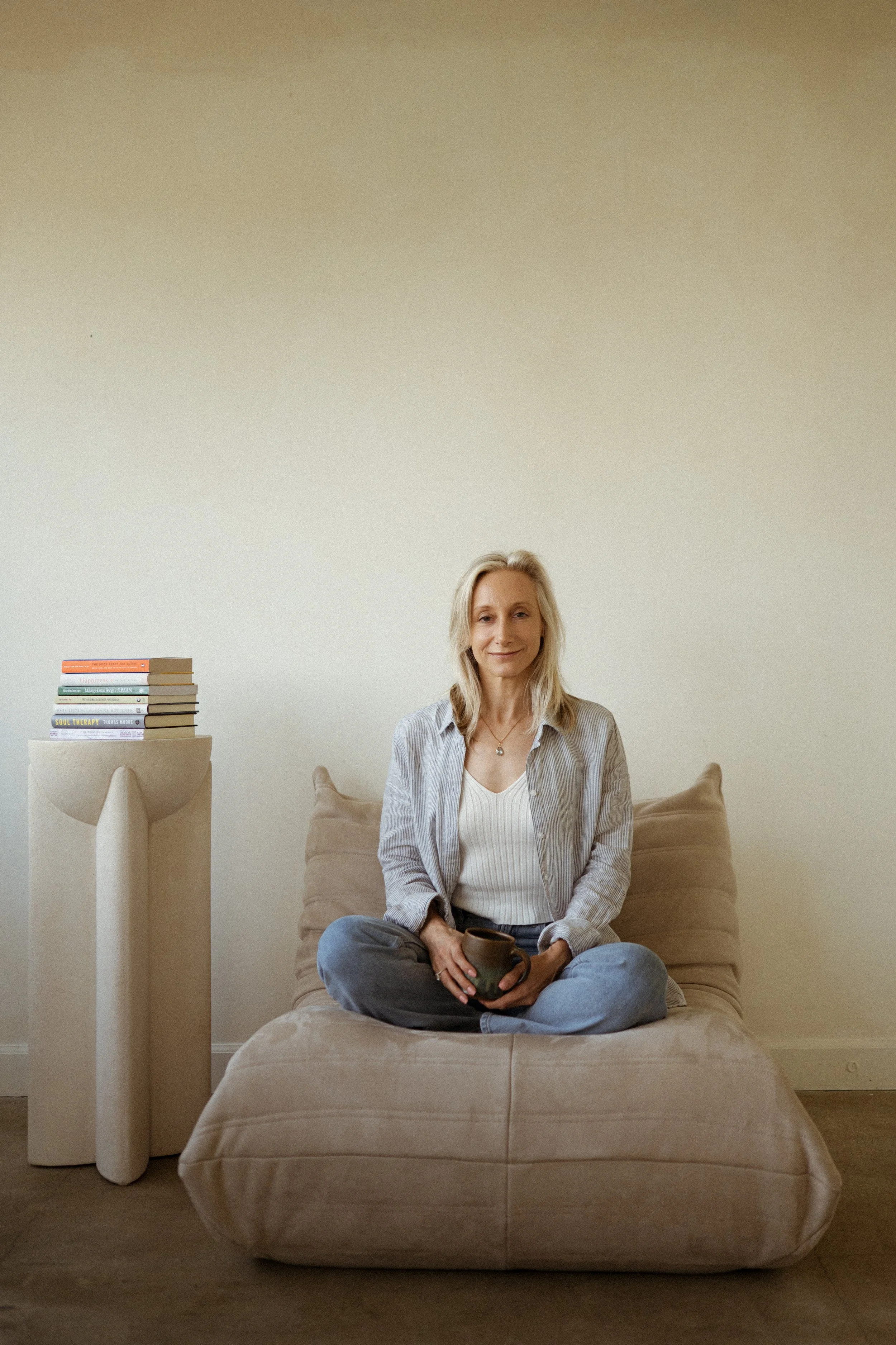 A woman sitting cross-legged on a beige sofa, holding a cup, with a stack of books on a side table next to her, in a minimalistic room with a plain wall.
