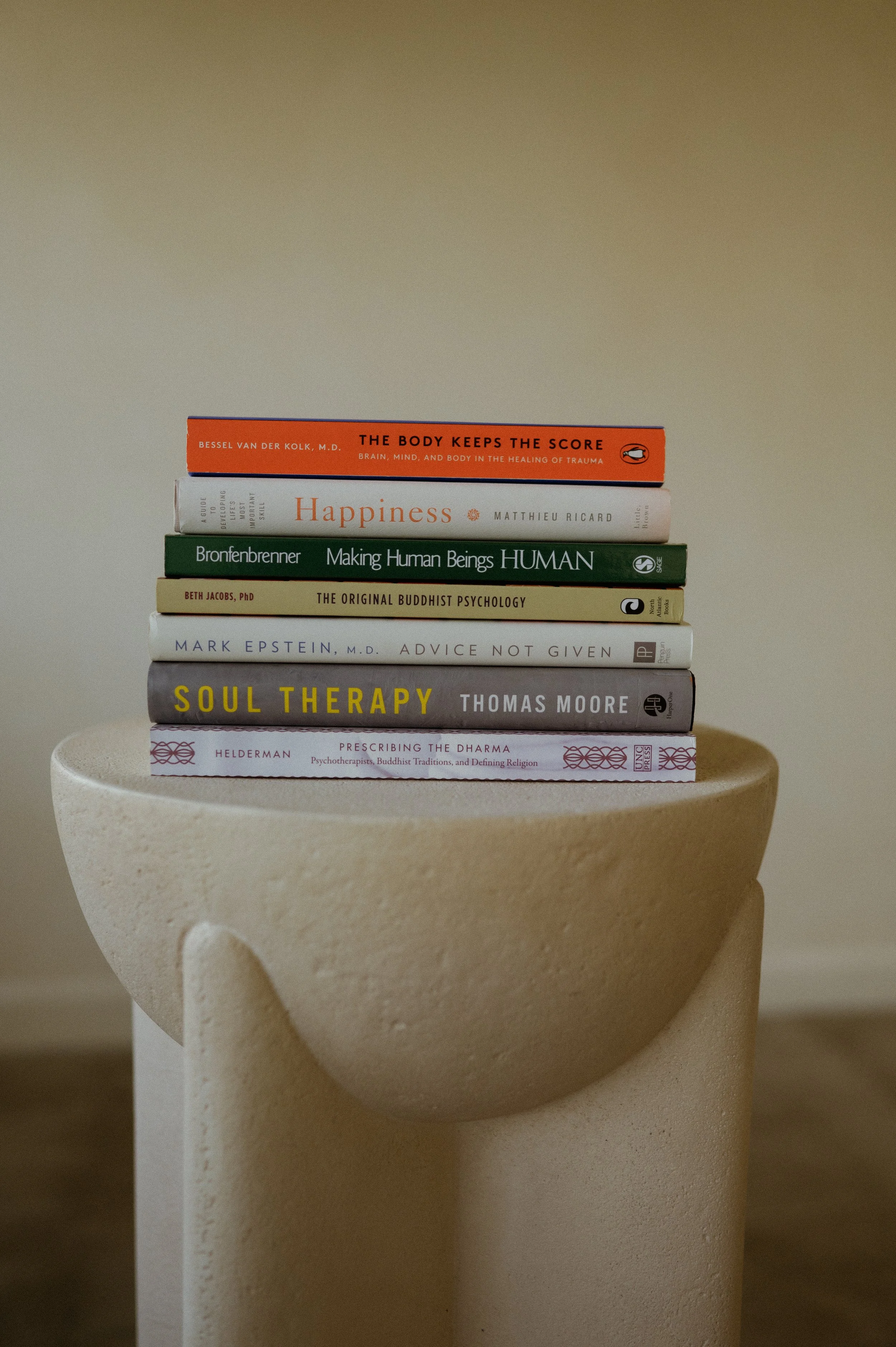 Stack of eight books on top of a beige, textured pedestal against a plain background.