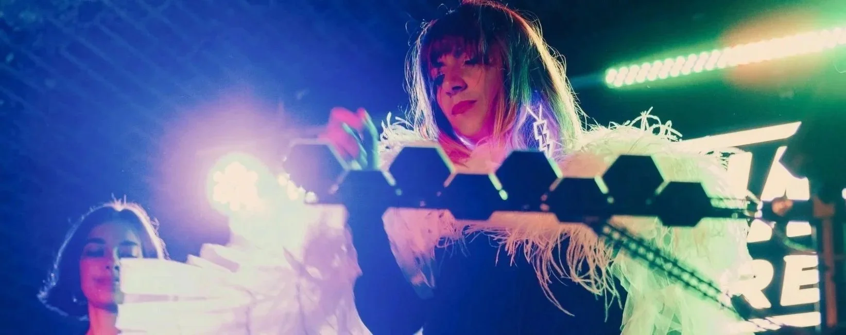 Two women on stage at a neon-lit event, one with colorful red hair and feathered outfit, with bright colorful lights in the background.