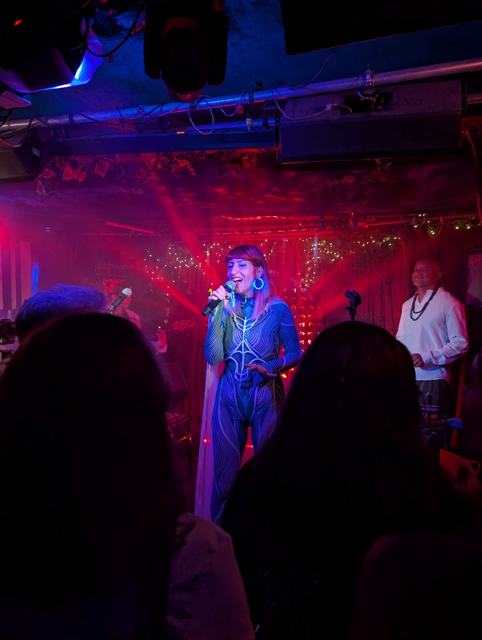 WUNDER BETA performing and singing into a microphone on a dimly lit stage and rays of red light behind her, with a man standing nearby and audience members in the foreground.