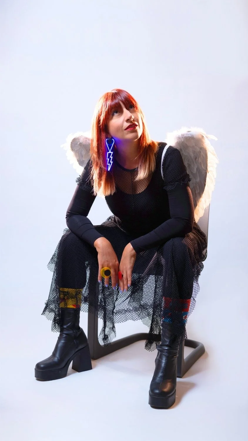 WUNDER BETA with red hair dressed in black, wearing angel wings and glowing blue earrings, seated on a simple chair against a plain white background.