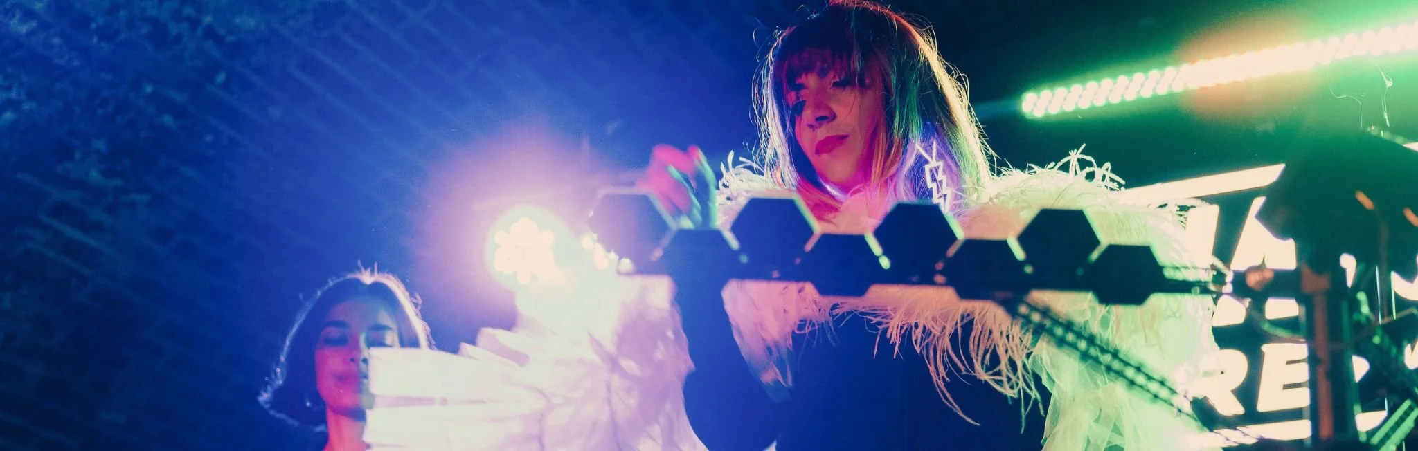 Two women playing electronic music with synthesizers at a concert, with rainbow-colored stage lighting