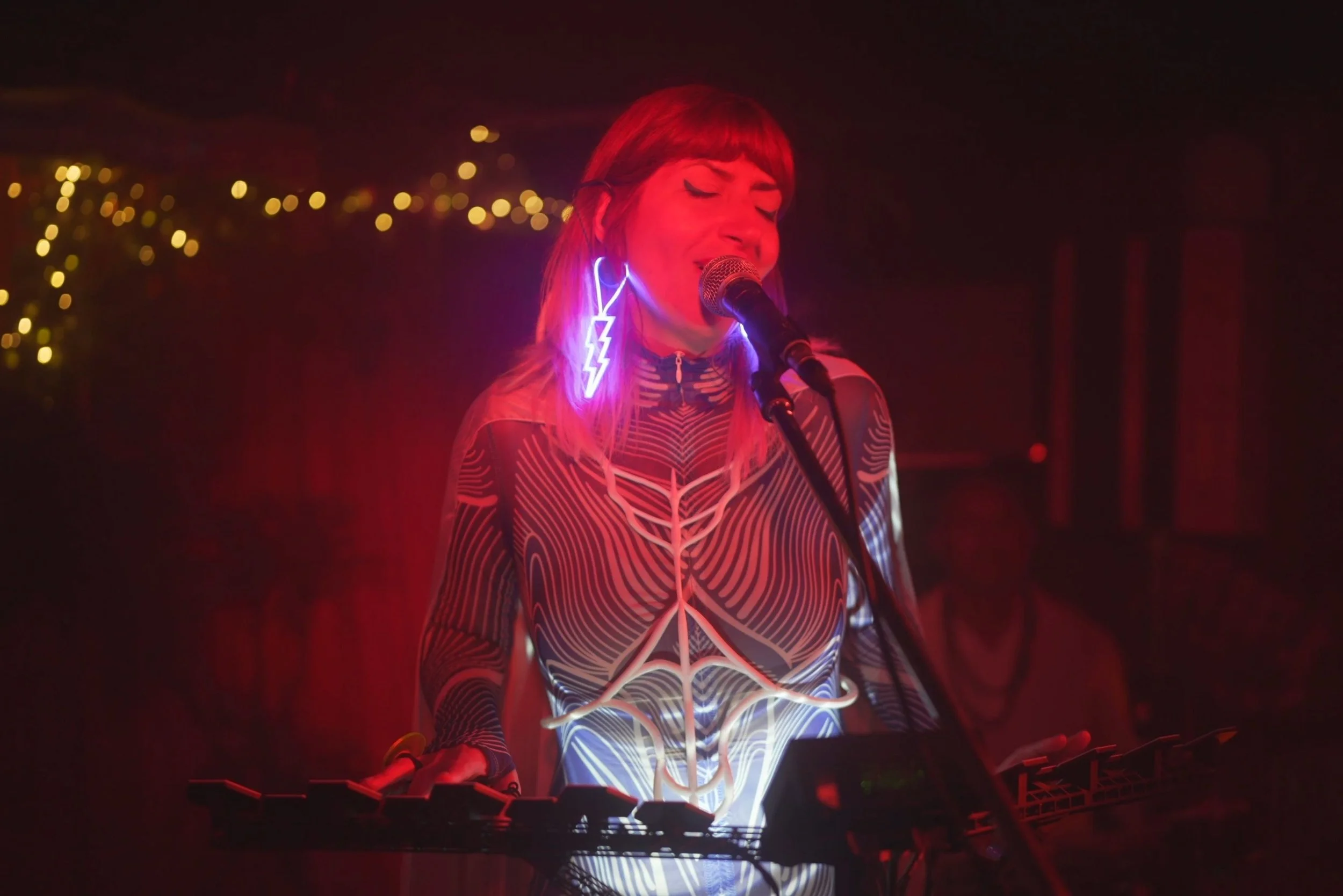 WUNDER BETA singing and playing MOrgana music instrument she made with neon lightning bolt earrings performing on stage, illuminated by red and purple lights, with a blurred background of string lights.
