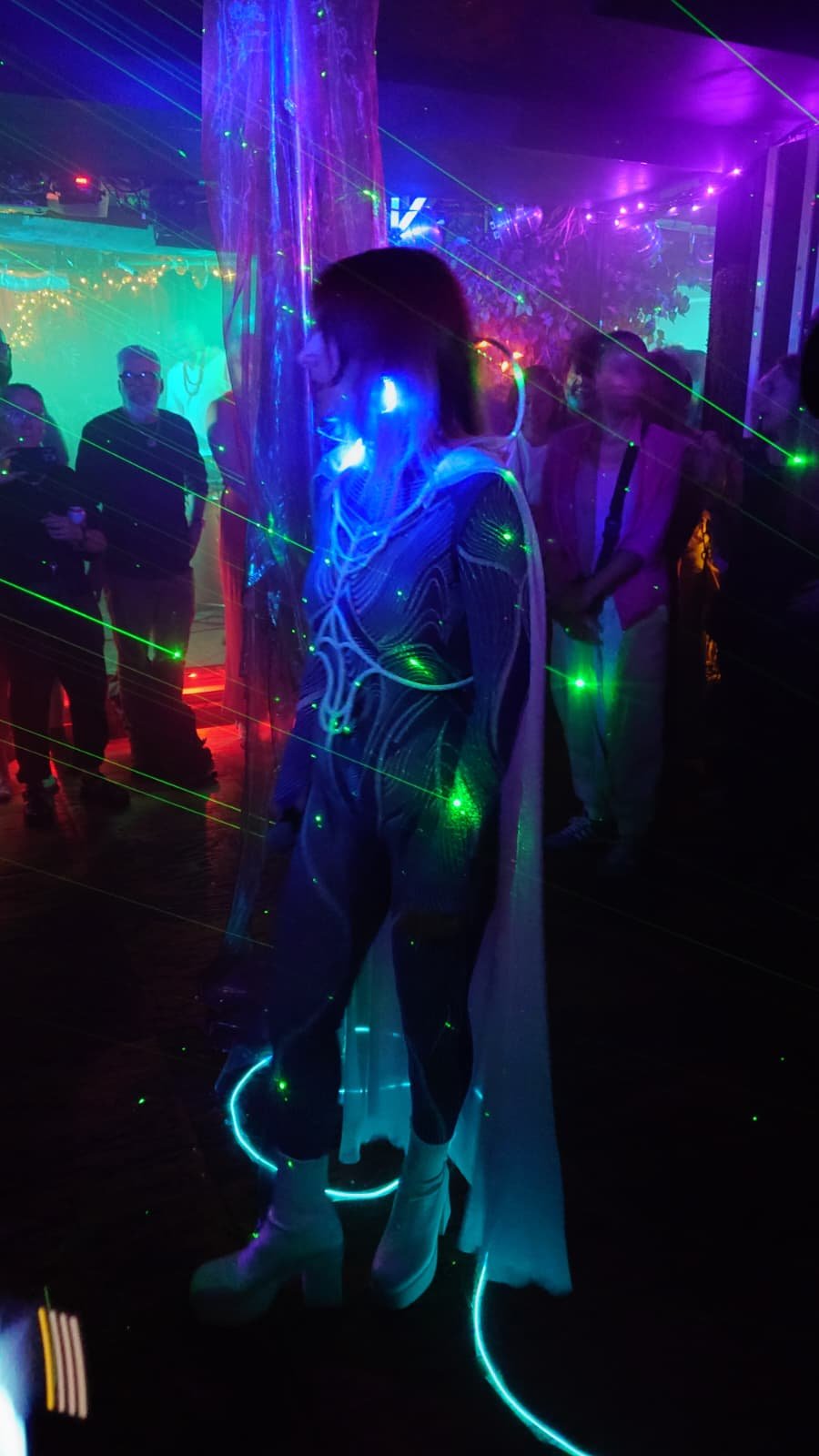 A person wearing LED light-up clothing and platform shoes at a colorful, neon-lit party with laser lights and other people in the background.