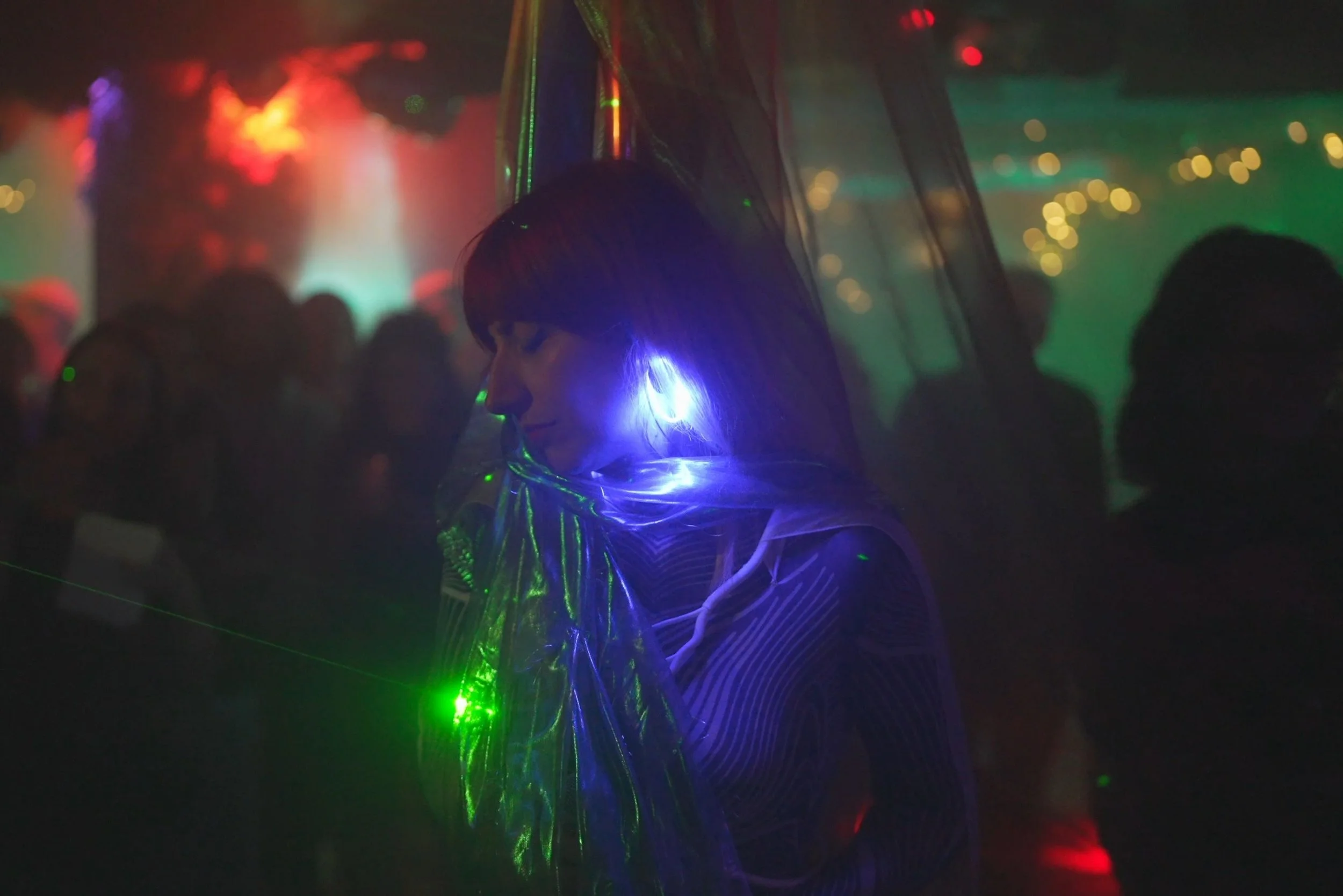 WUNDER BETA performing with closed eyes in a dark room illuminated by colorful lights and laser reflections, with a blurred crowd in the background.