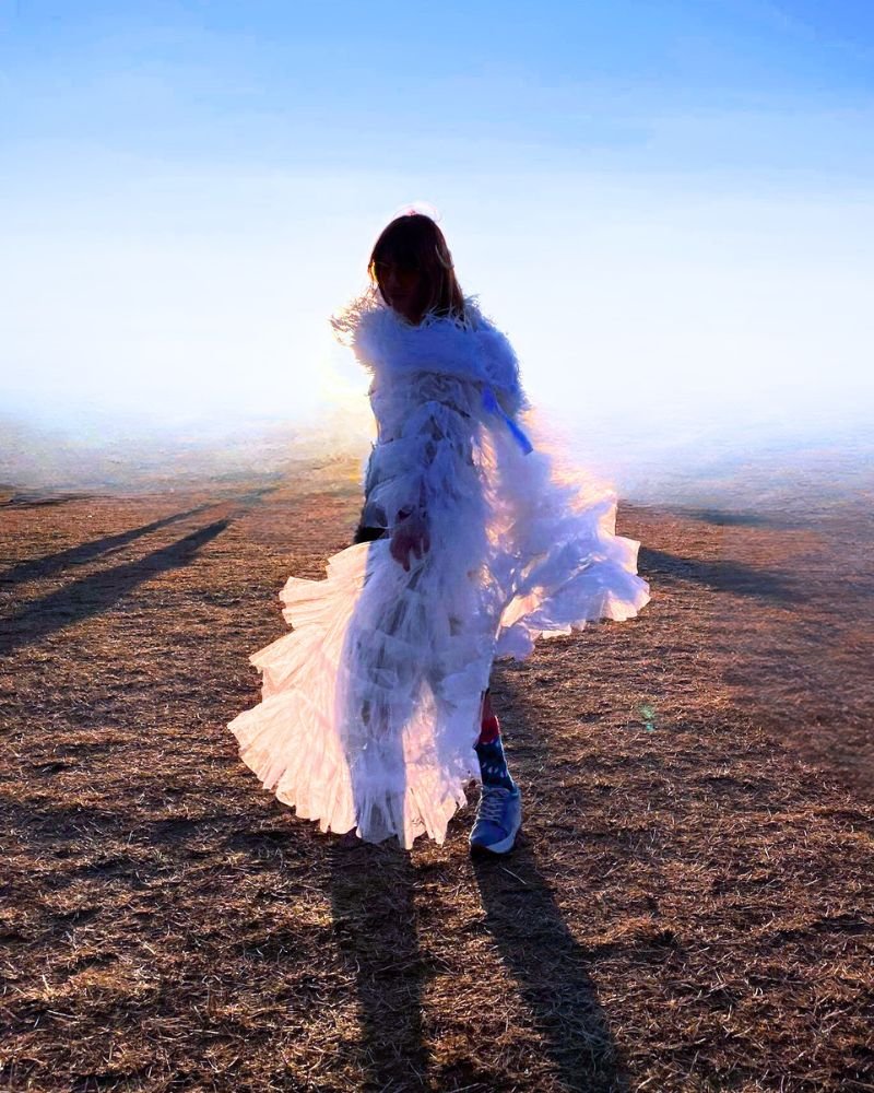 A person dressed in a flowing white gown standing in a desolate landscape during sunset or sunrise, with long shadows cast on the ground and the sky glowing with a soft light.