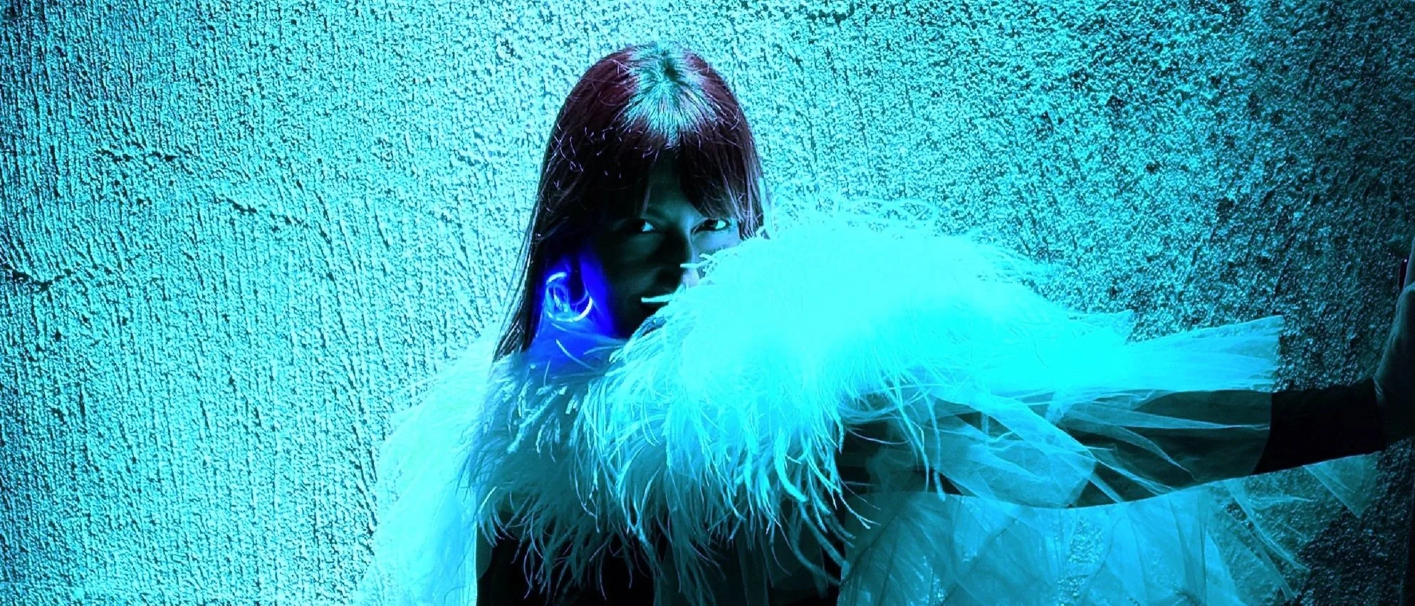 Person wearing a black outfit and large feathered white garment, illuminated with blue light, standing against a textured wall.