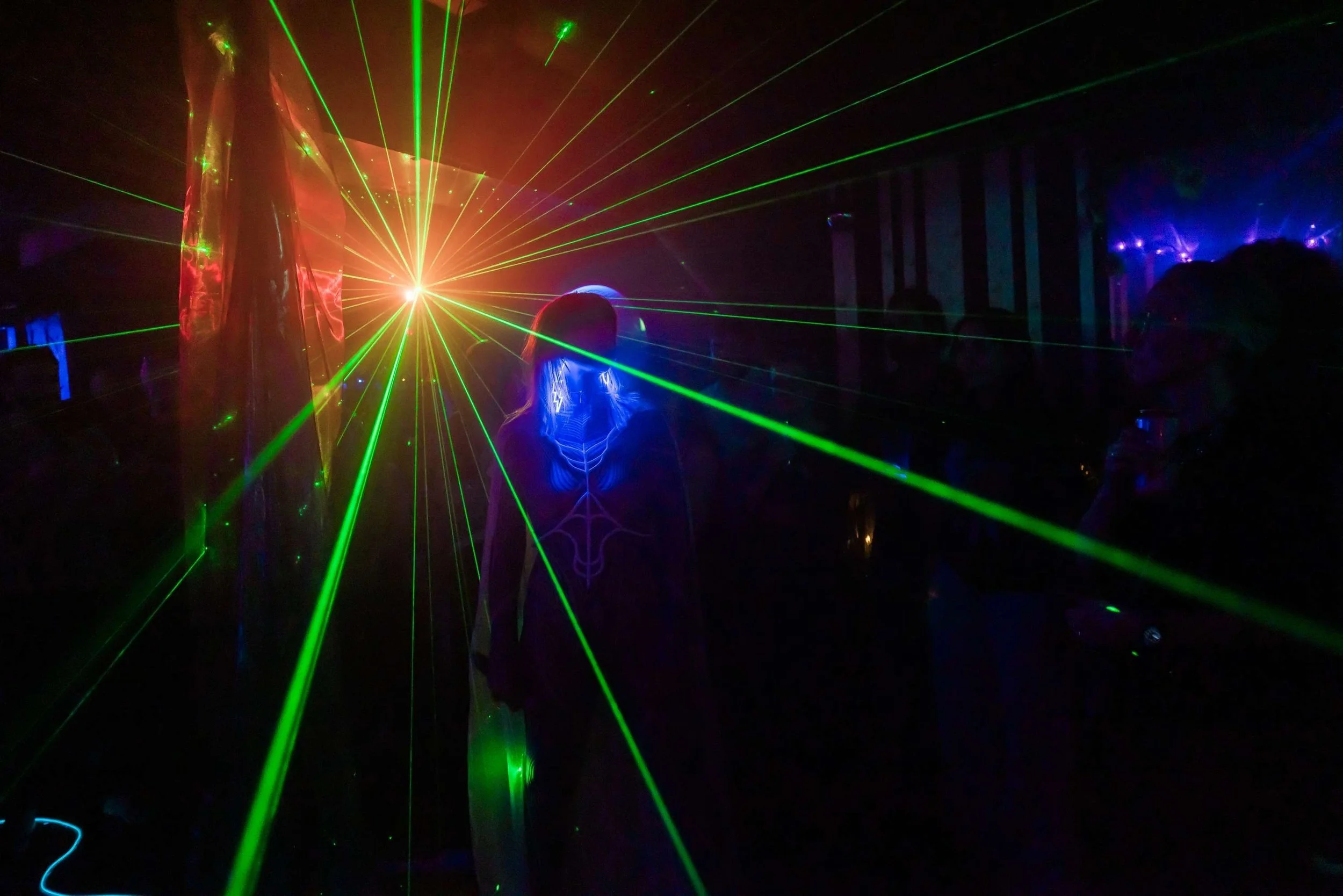 WUNDER BETA performing live at Live Transmission Show in London and a group of people in a dark room with laser lights and projections, standing and enjoying a laser show.