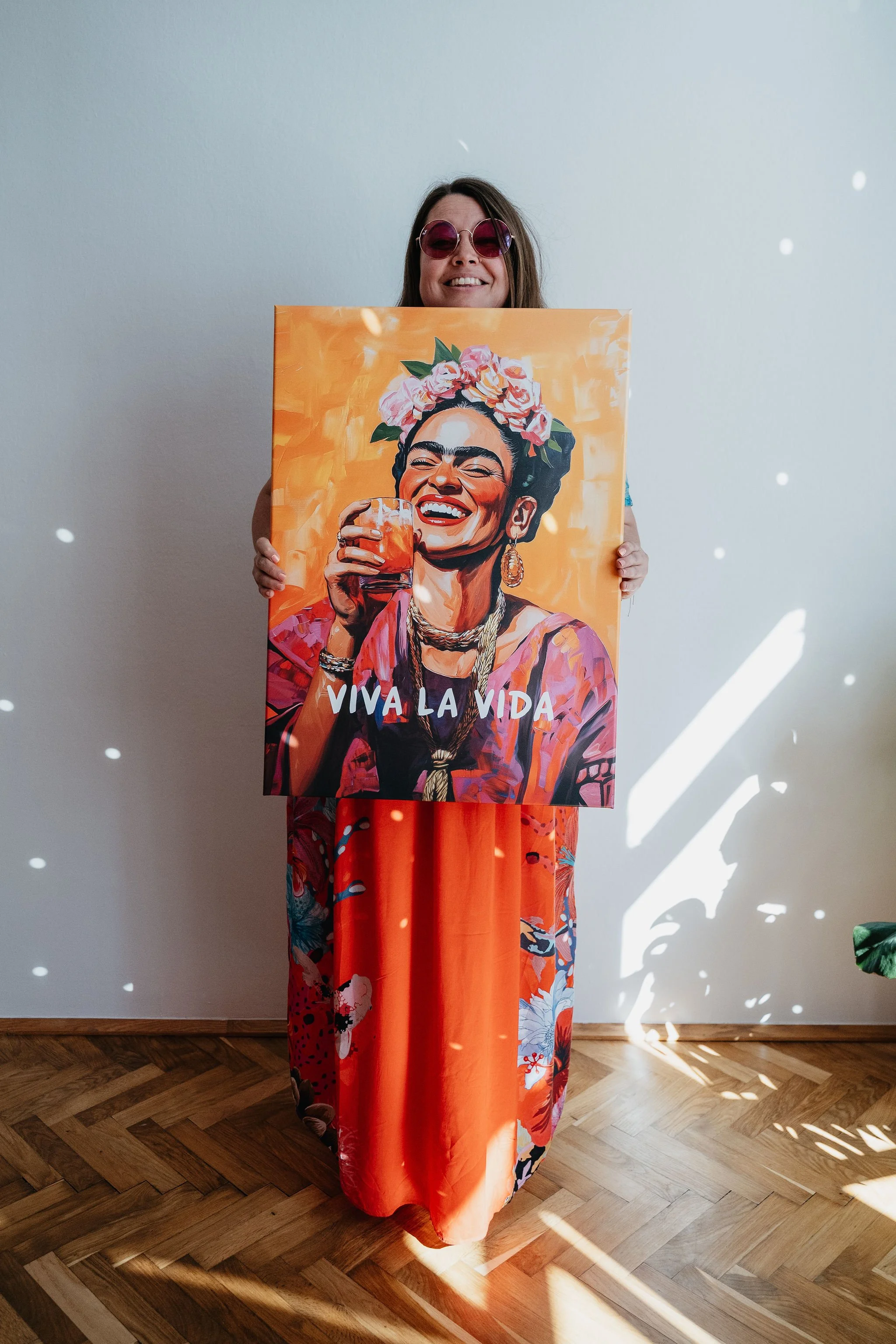 Lächelnde Frau mit Sonnenbrille hält ein farbenfrohes „Viva la Vida“-Gemälde mit Blumenkranz und Cocktailglas.