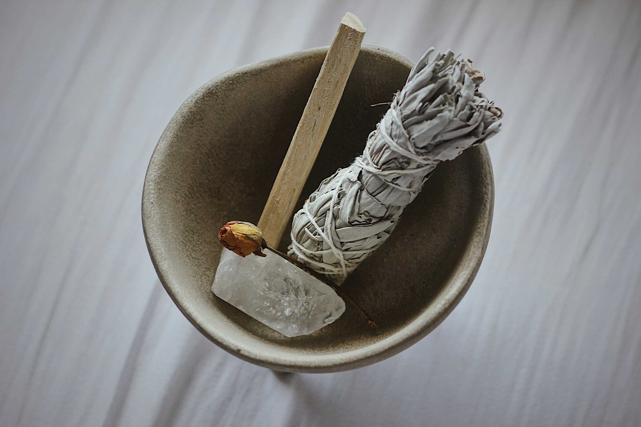 A bundle of smudging sage wrapped in white paper, a wooden stick, and a dried rose are in a small beige bowl with a piece of clear quartz inside, on a light wood surface.
