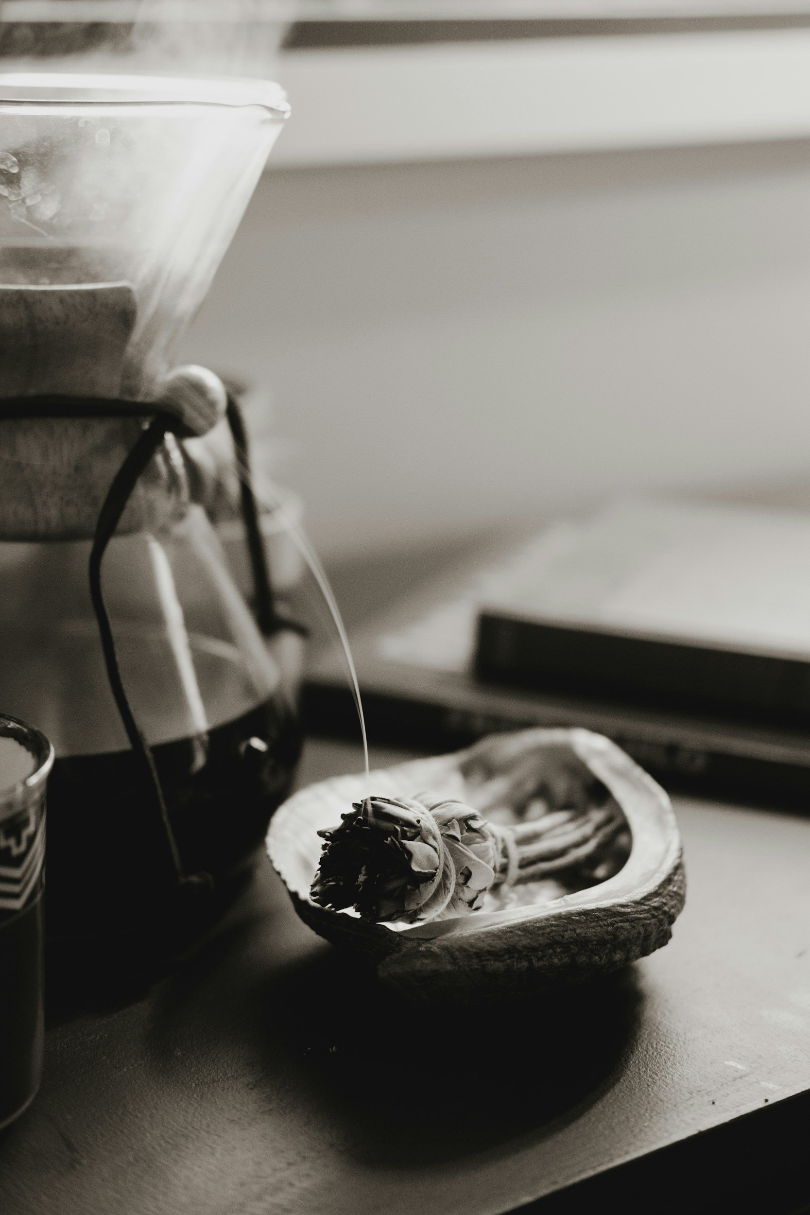 A black and white photo of a small ceramic bowl with a dried flower inside, placed on a table near a jar of liquid, possibly tea or coffee, with a blurred closed book or notebook in the background.