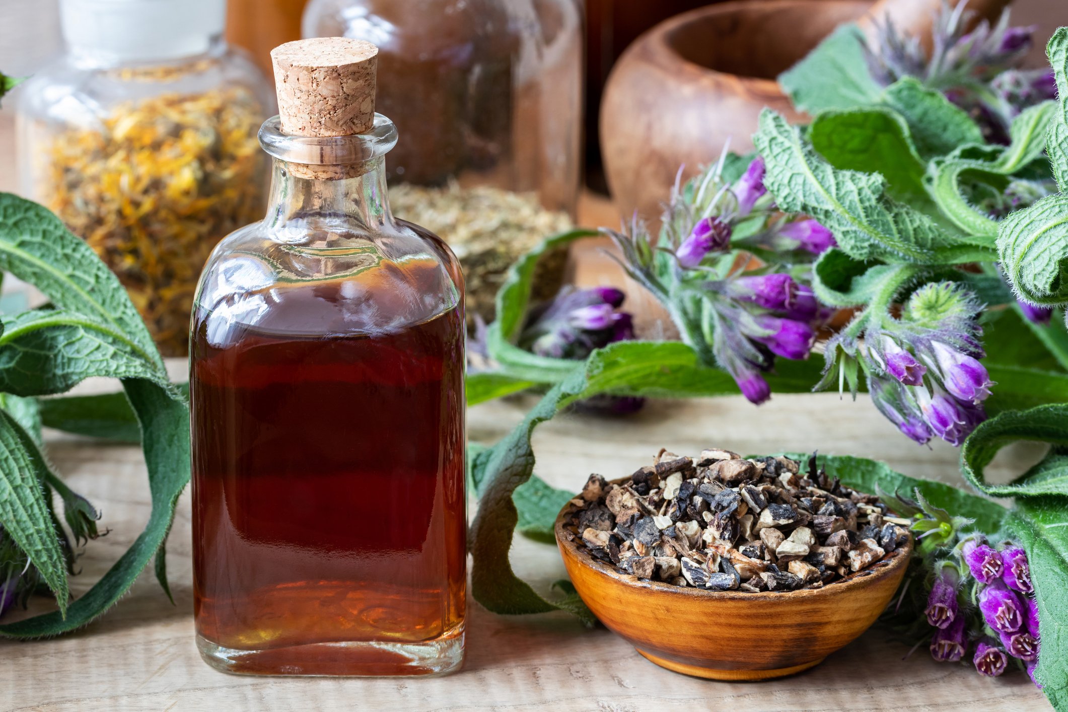 Herbal tincture in a glass bottle with a cork next to purple flowering herbs, dried herbs, and a bowl of chopped herbs on a wooden surface.