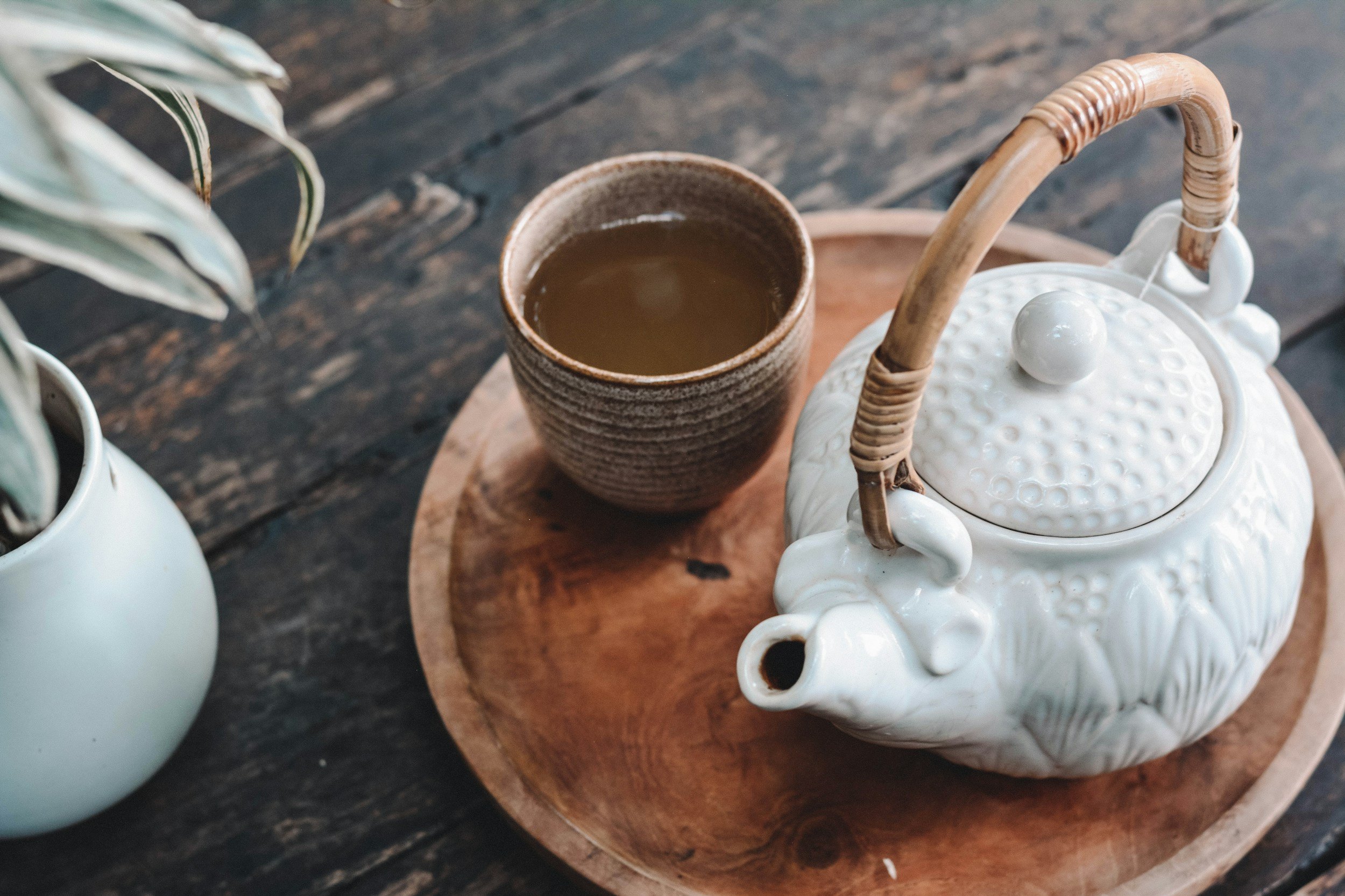 White ceramic teapot with bamboo handle, a brown ceramic cup filled with tea, and a white vase with green and white variegated leaves on a wooden tray and table.