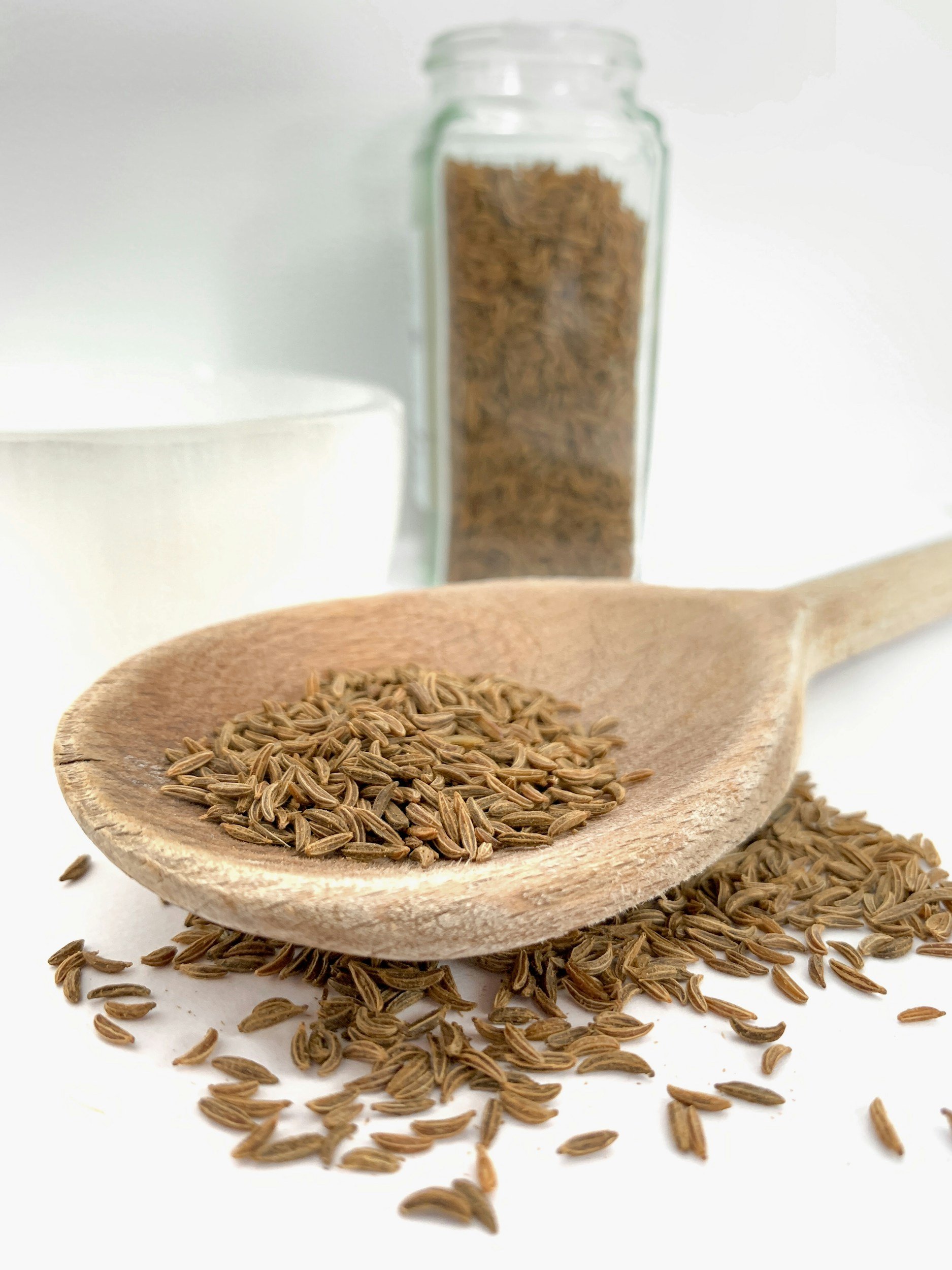 Wooden spoon filled with cumin seeds, spilled on a white surface with a jar of cumin seeds and a bowl in the background.