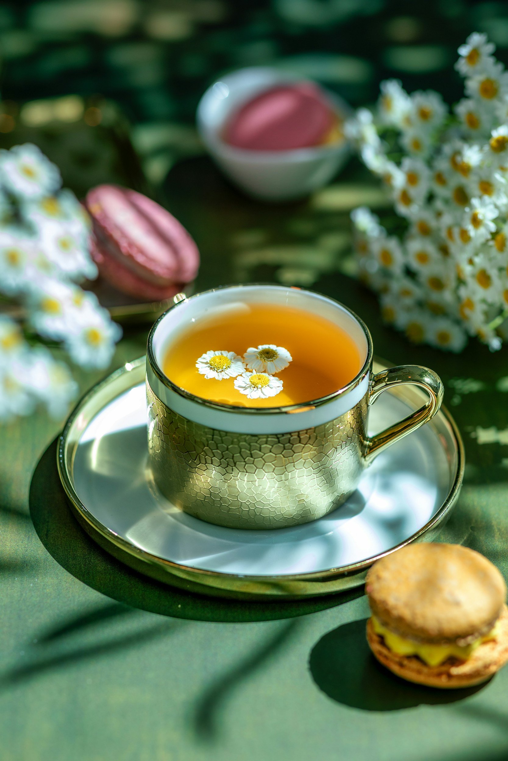 A gold-rimmed teacup filled with tea, garnished with small white daisies, sits on a silver tray on a green surface. In the background, there are pink macarons, a small bowl of pink cream, and white daisies.