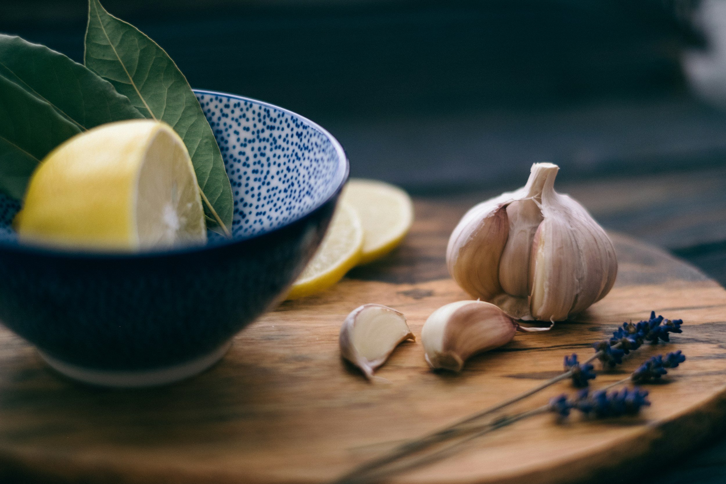 A garlic bulb with loose cloves, lemon slices in a blue patterned bowl, and sprigs of lavender on a wooden surface.