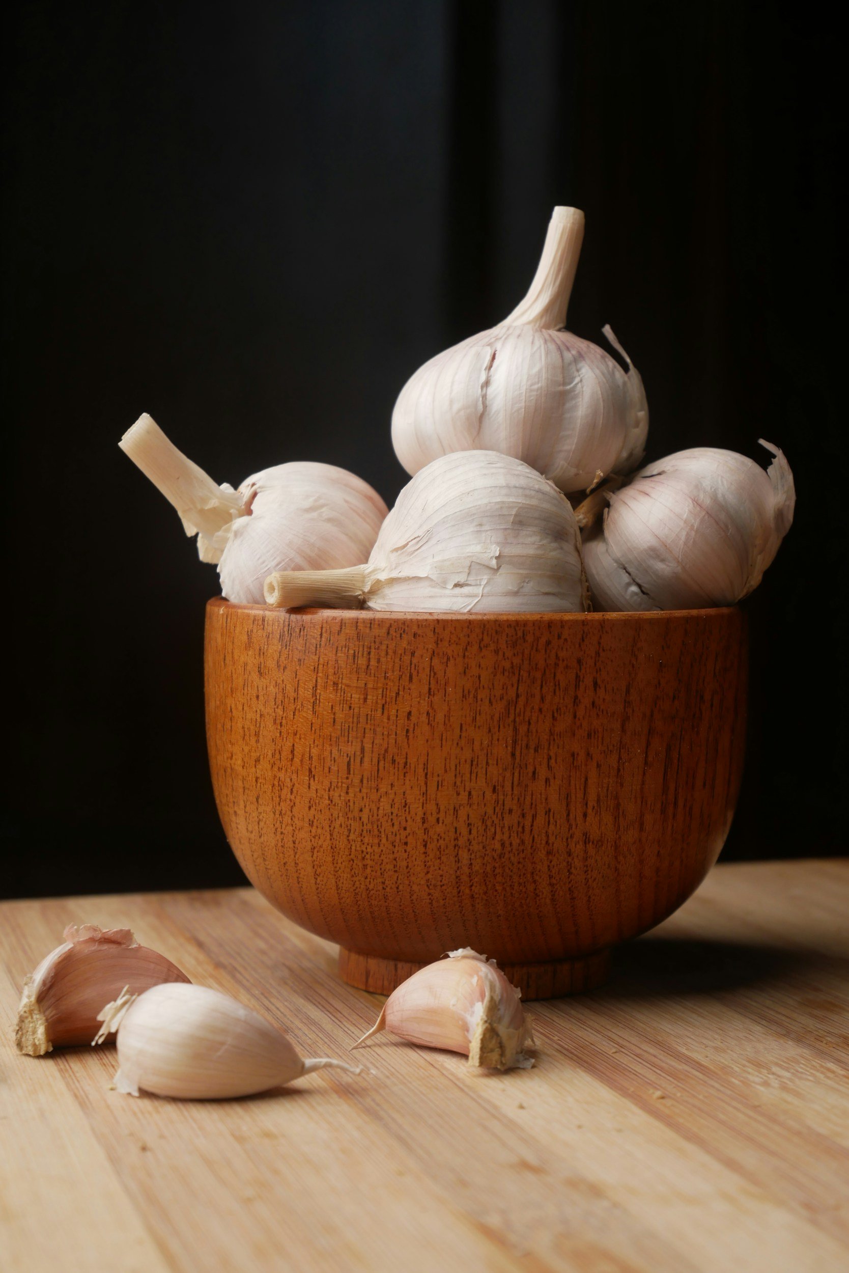 A wooden bowl filled with several heads of garlic set on a wooden surface with loose garlic cloves in the foreground, against a dark background.