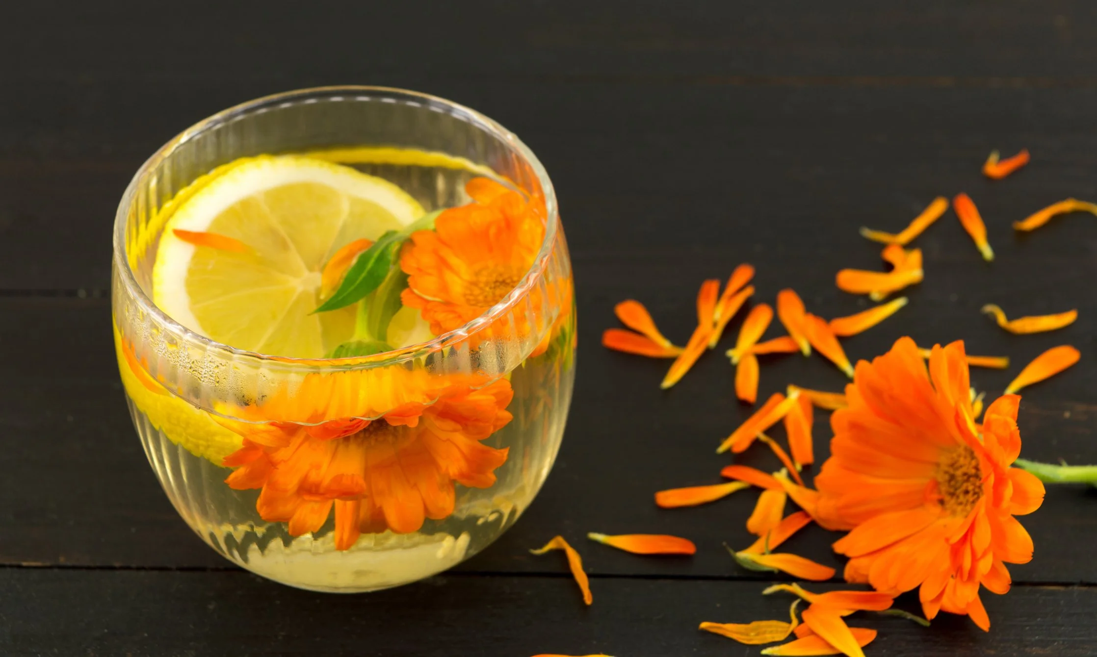 Glass of lemon water garnished with orange flowers and lemon slices, with scattered orange flower petals on a dark wooden surface.