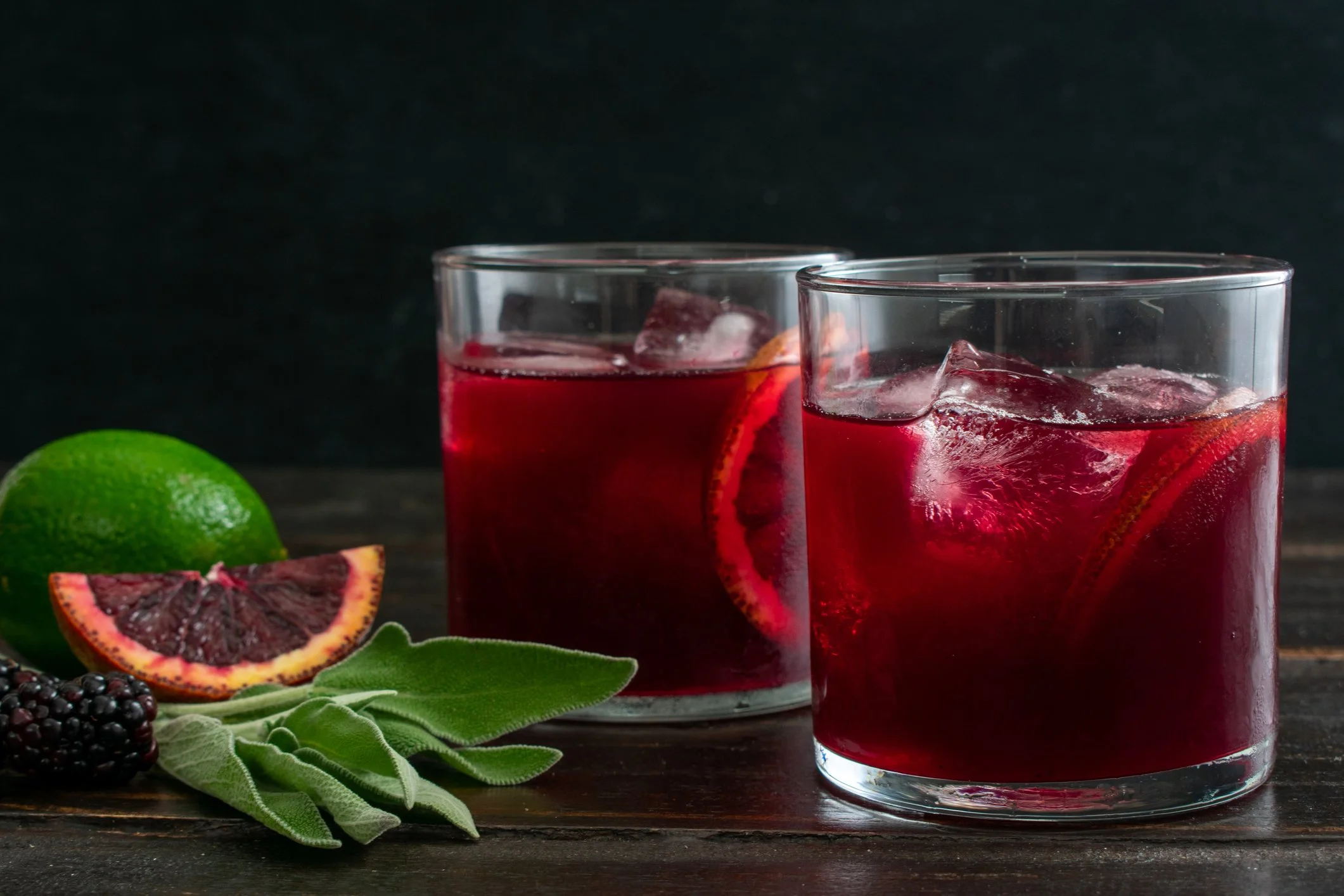 Two glass tumblers with blackberry and sage cocktail with sage and blood orange garnish.