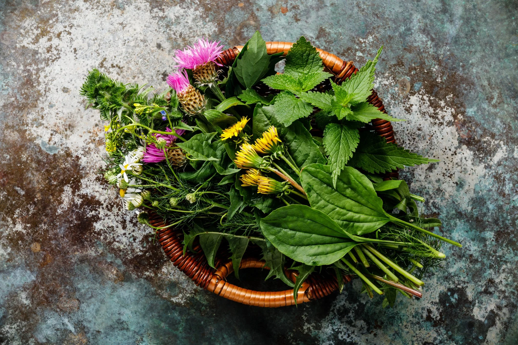 Basket filled with various fresh wildflowers and green leaves, placed on a textured, weathered surface.