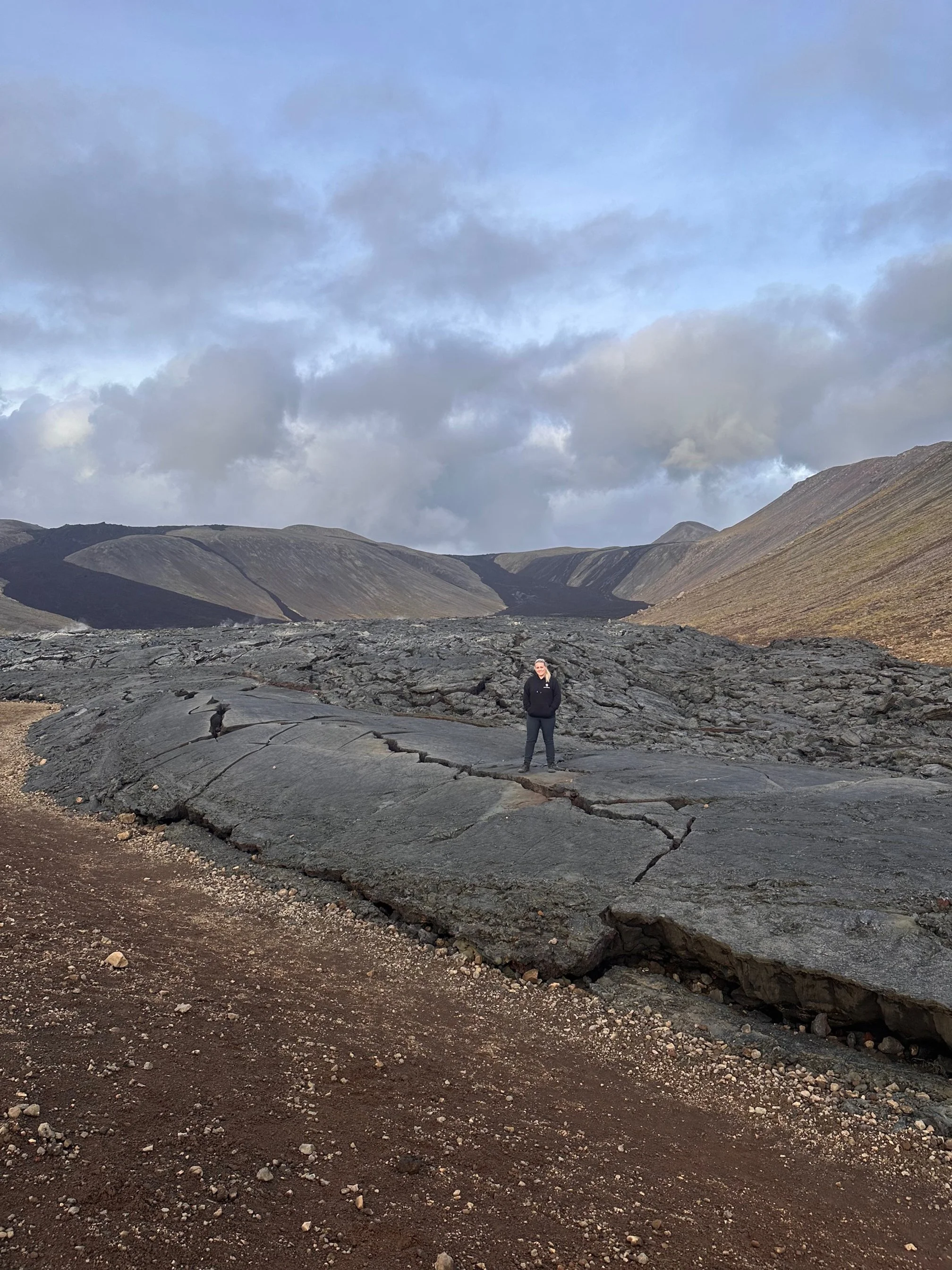 Owner of website standing on solidified lava field, with the harden lava cascading down the hills in the background. 