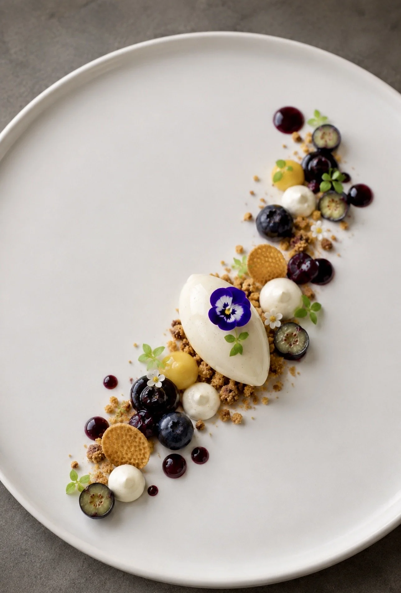A modern dessert with blueberries, yellow fruit, white dollops, edible flowers, and microgreens arranged on a white oval plate.