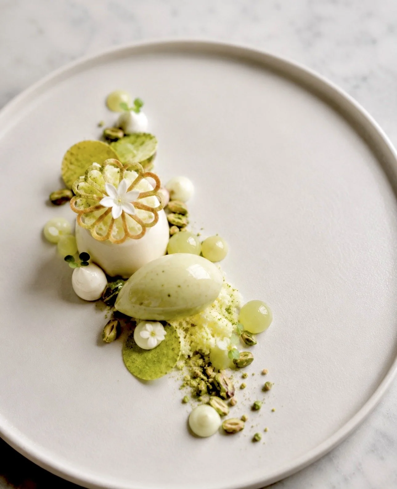 Elegant dessert with white chocolate or mousse, garnished with edible flowers, pistachio pieces, and green chocolate discs on a white plate.