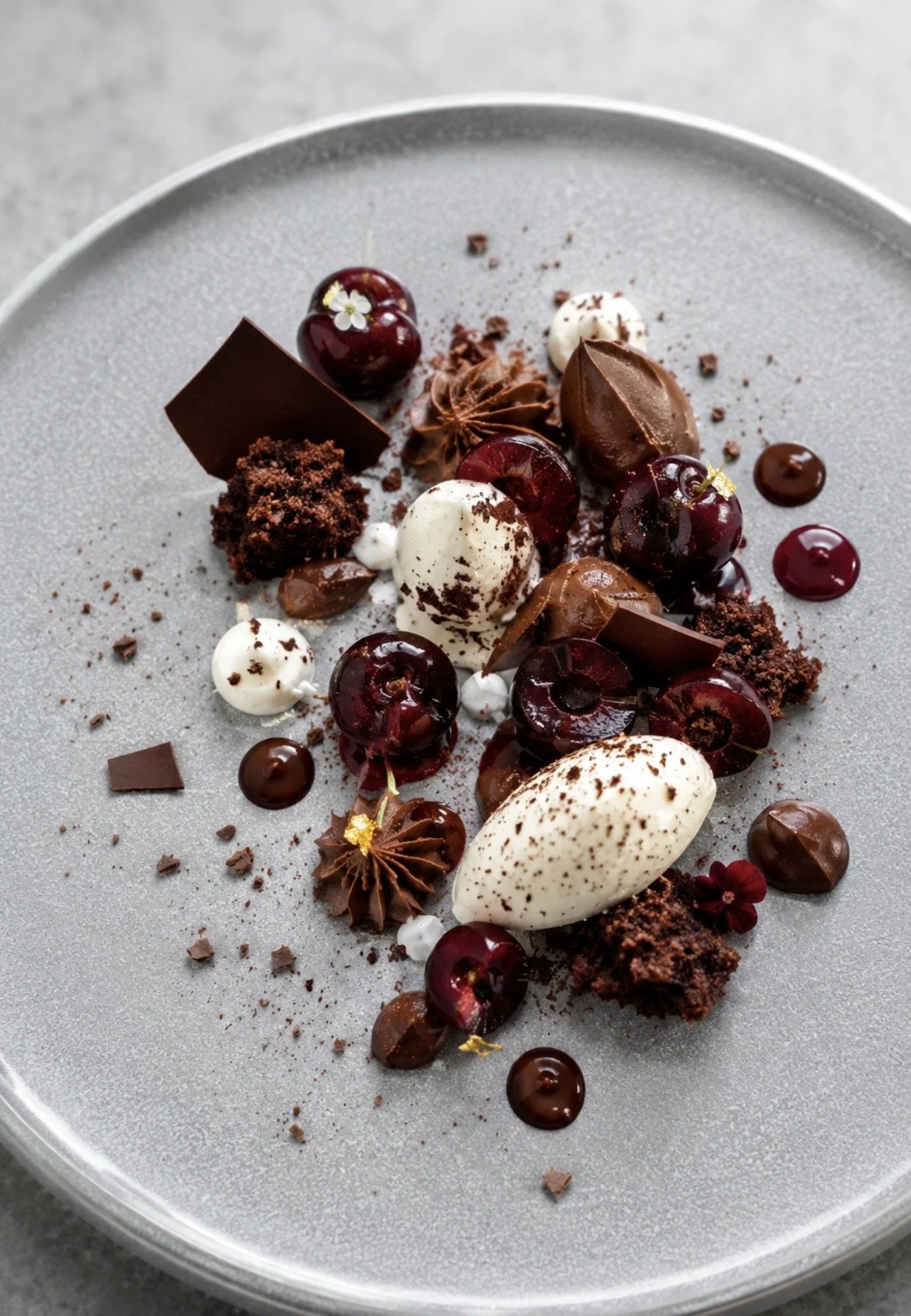Elegant dessert plate with cherry garnishes, chocolate curls, cream, and edible flowers on a gray ceramic dish.