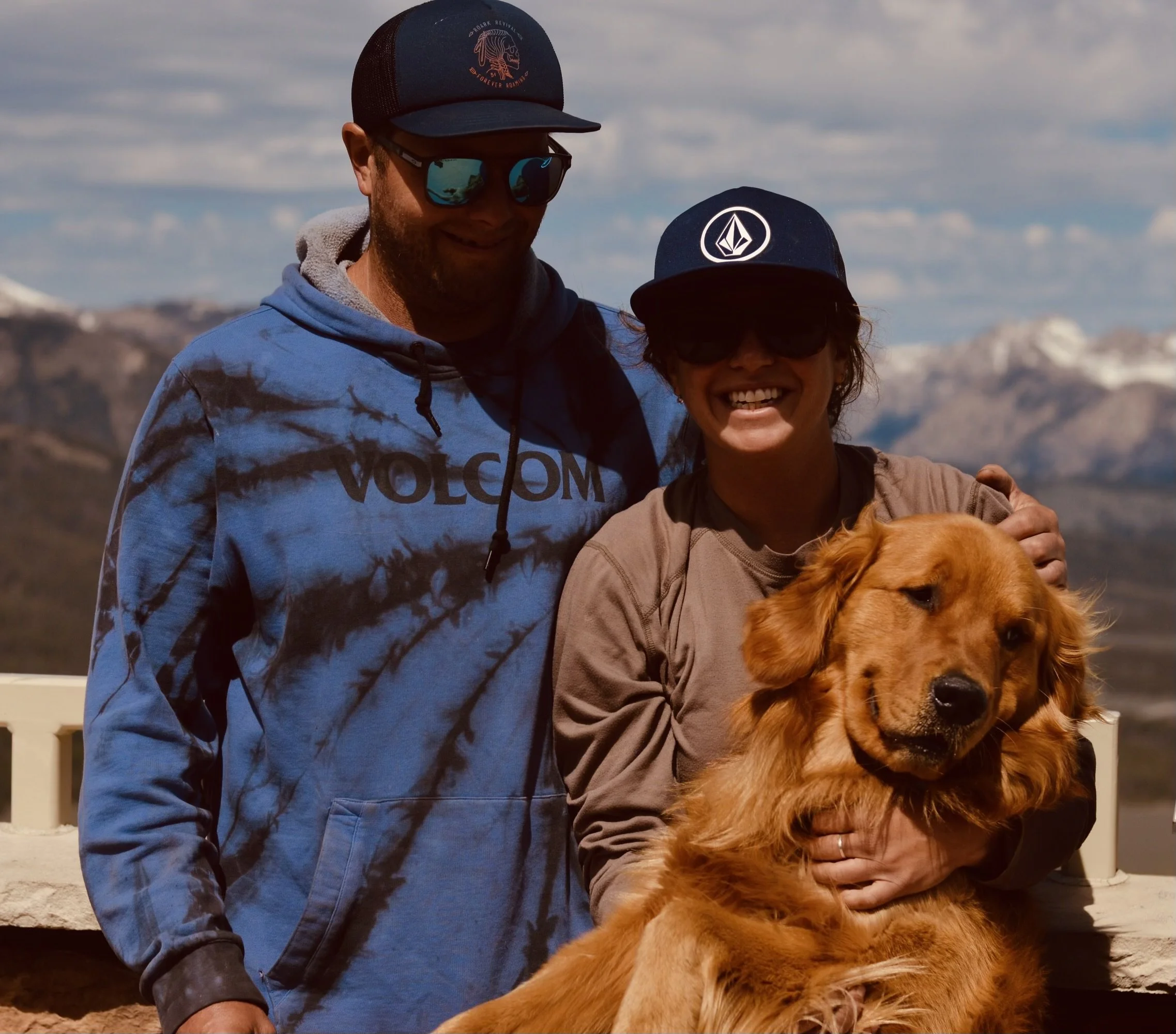 A couple outdoors with a golden retriever. The man is wearing a blue hoodie and baseball cap, and the woman is wearing dark sunglasses and a cap with a logo. They are smiling, and the dog is being held lovingly by the woman. A mountainous landscape is visible in the background.