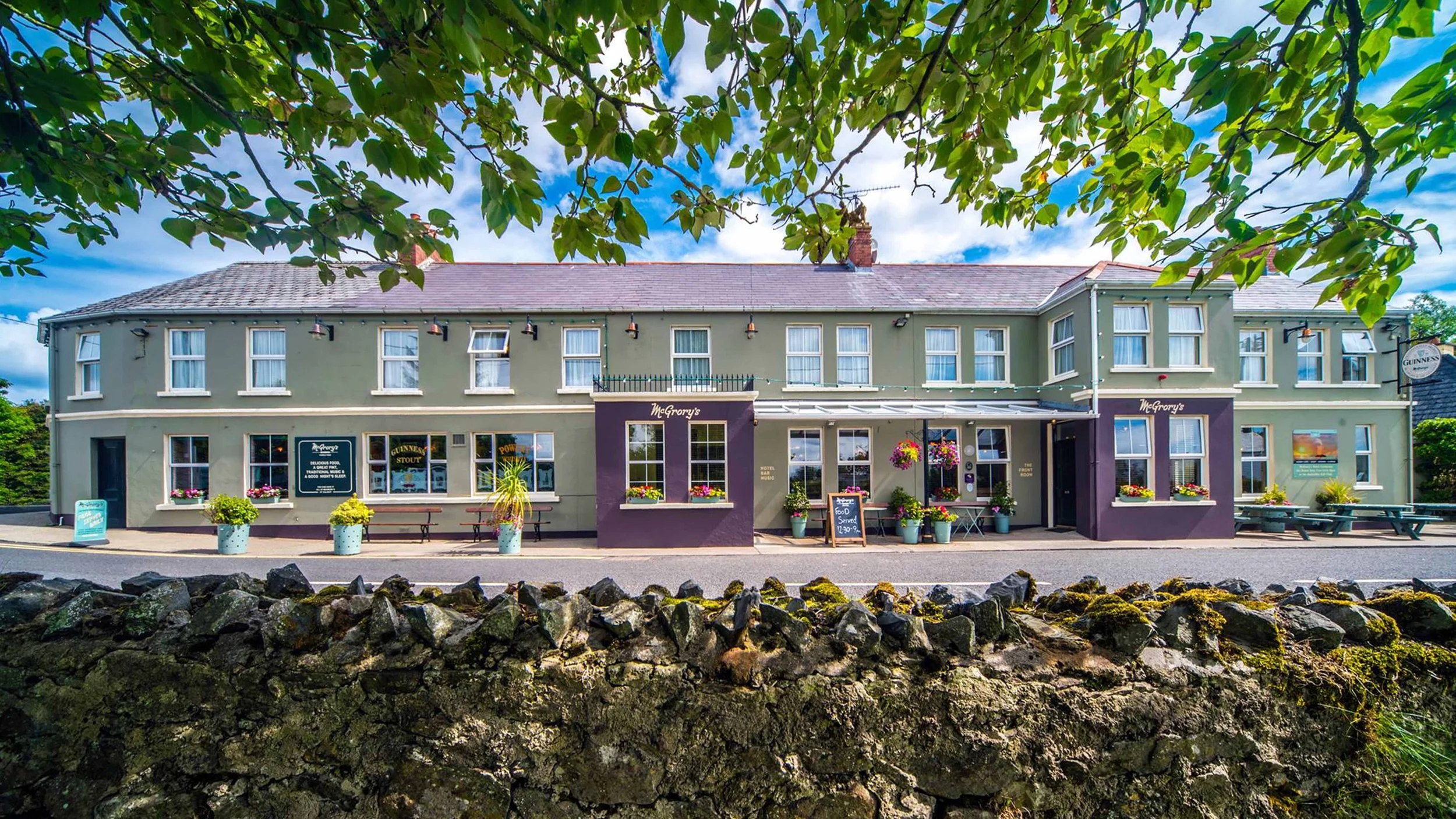 Front view of a two-story pub called McGorry's, with pastel green facade, purple accents, and flower pots outside, under a leafy tree with a partly cloudy sky.