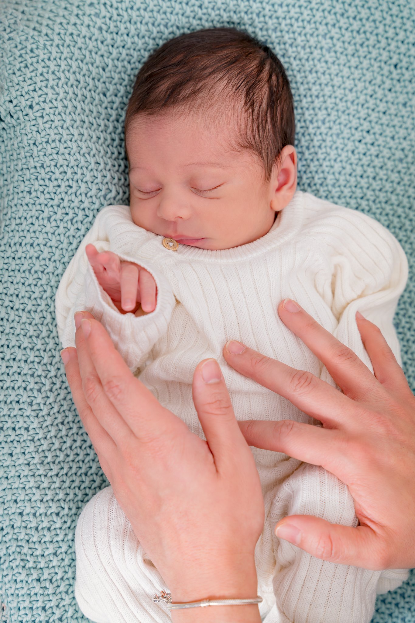 Nourisson habillé en blanc sur une couverture tricotée turquoise pastel  est bercé par les mains de sa mère 