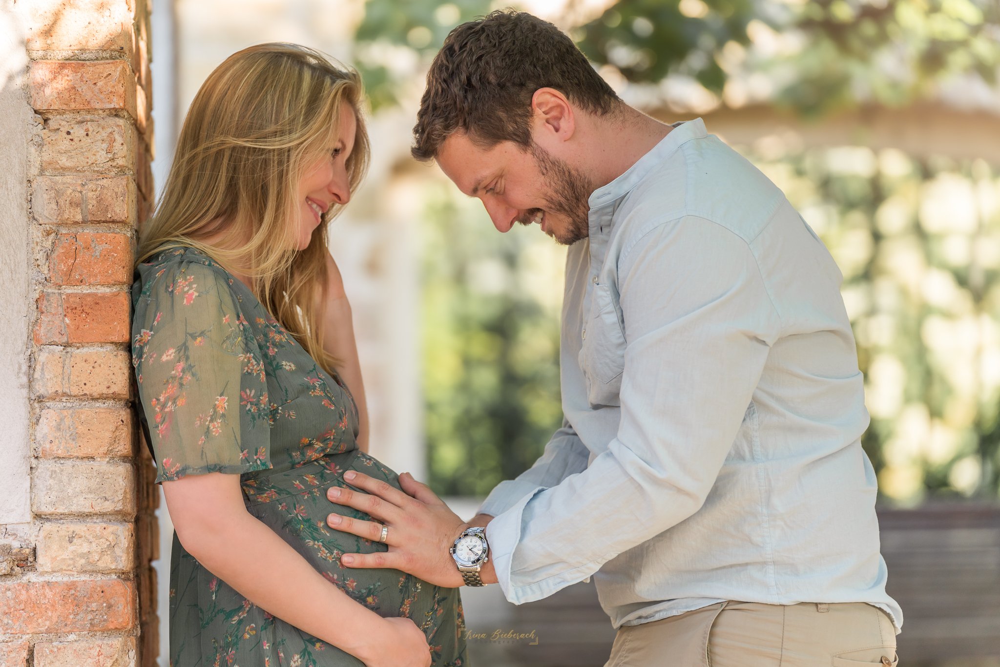 Un futur papa touche le ventre rond de sa femme qui le regarde souriante dans le parc de l'Ile Saint Germain