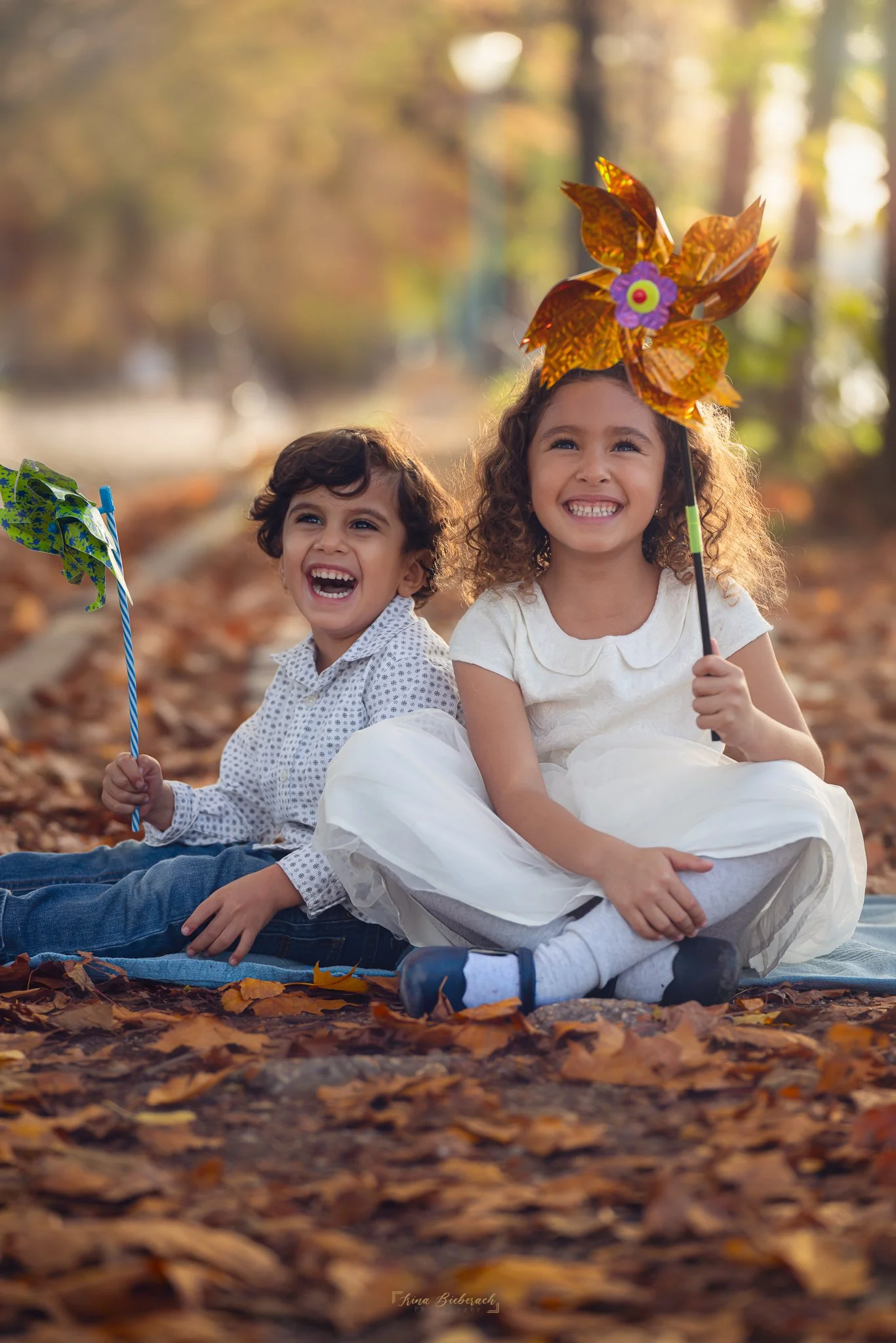 Petit garcon et petite fille souriants assis sur des feuilles oranges sèches en tenant de moulin de vents dans la main pendant un coucher de soleil d'automne dans l’allée des Cygnes à Paris