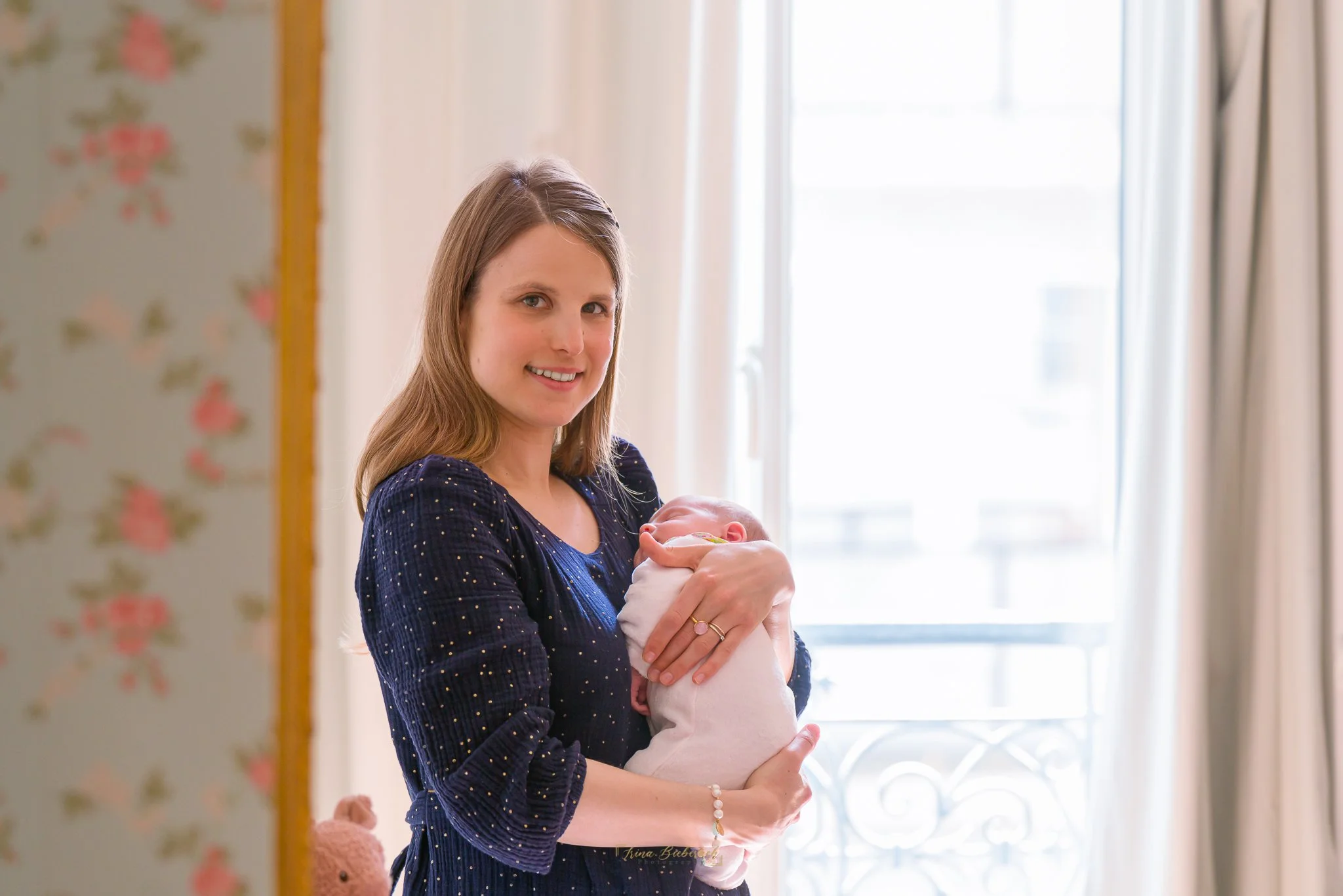 Nouvelle maman porte son nourrisson habillée en body rose dans une chambre illuminée par la lumière naturelle de la fenêtre 