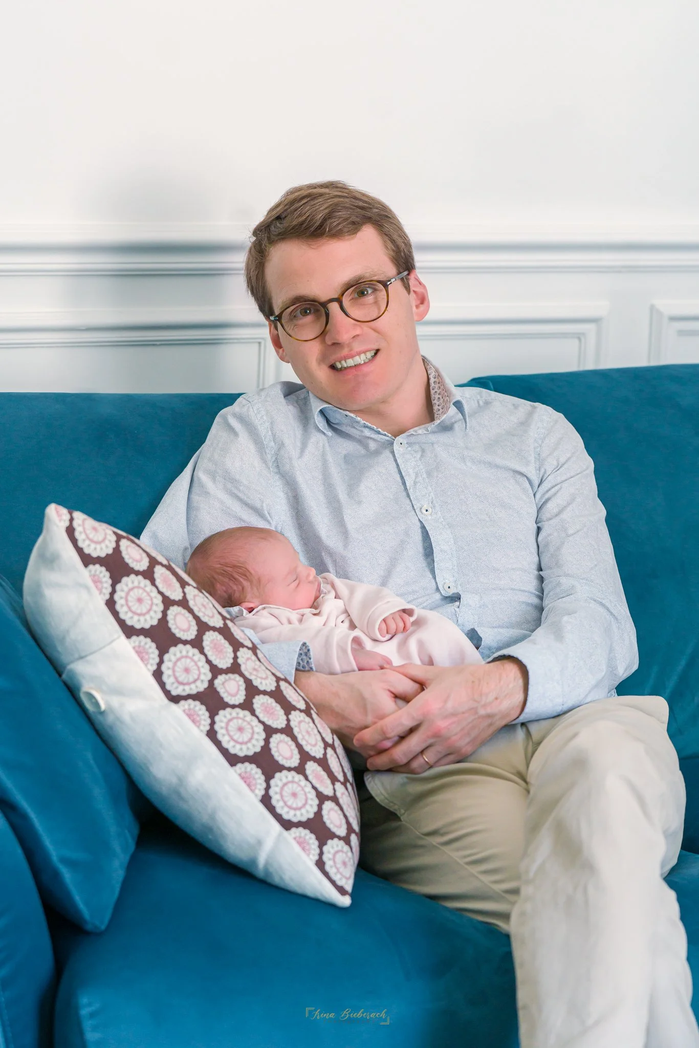 Jeune papa souriant assis sur un canapé d’appartement porte son bébé qui dort 