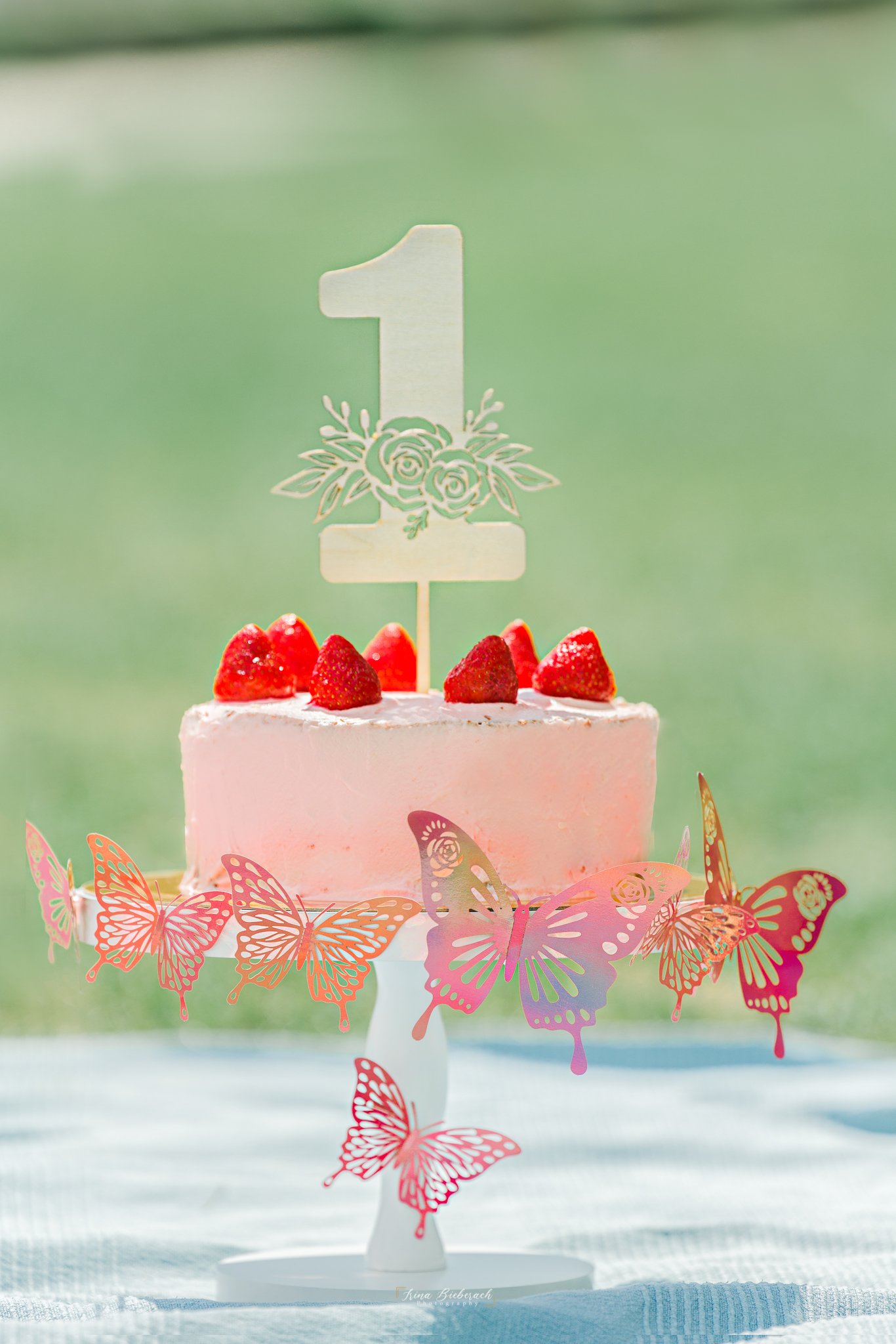 Gâteau d'anniversaire rose avec des fraises, sur un support rond de bois blanc et décoré avec des papillons roses, et le chiffre un en bois
