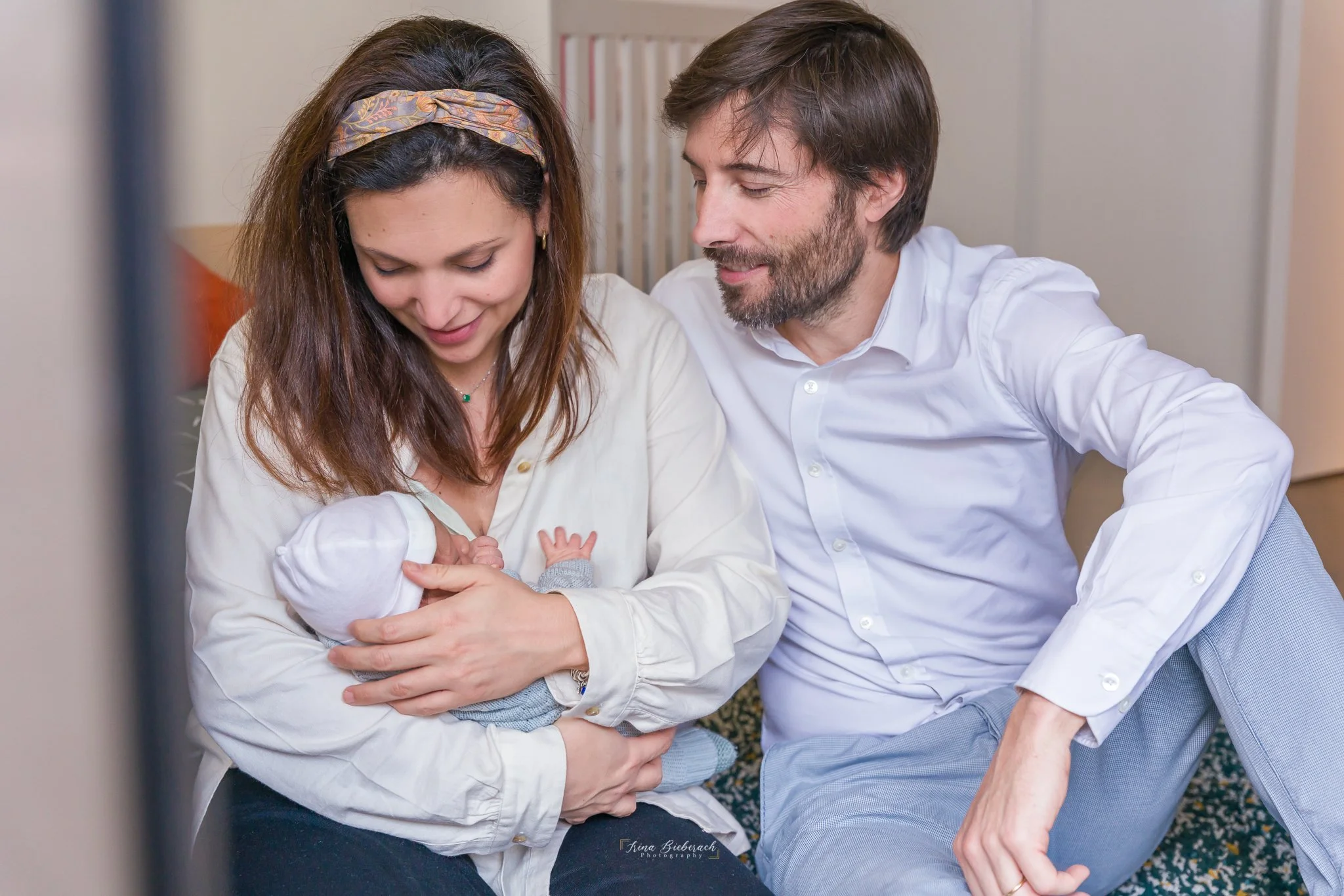 Maman souriante et papa souriant regardent leur bébé en bras de sa maman 