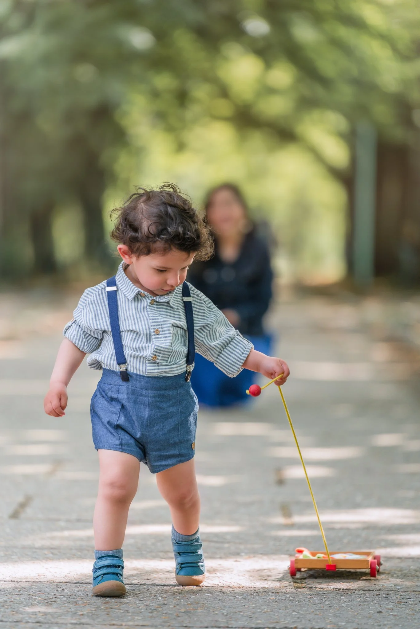 Petit garçon jouet avec un jouet en bois pendant que sa mère le regarde de loin dans un parc Parisien 