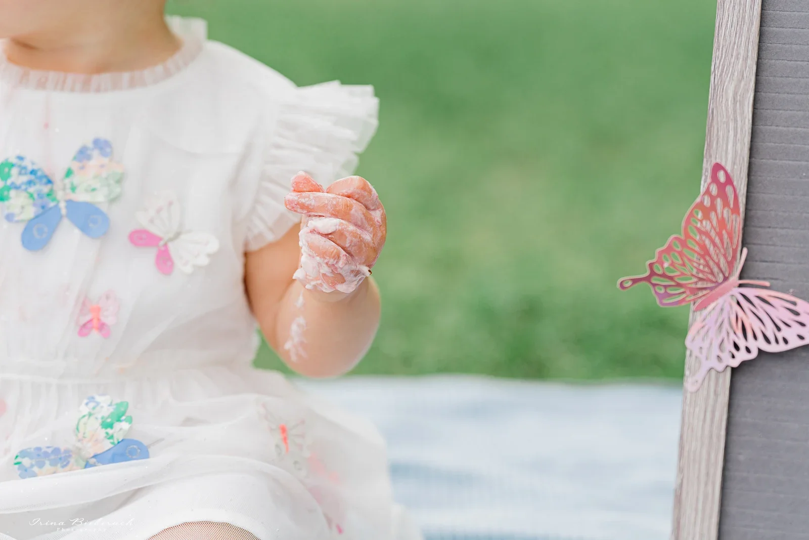 Détail de la main d'un enfant de 1 an couverte de crème de gâteau rose, avec un papillon de décoration à côté.